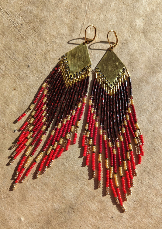 Moon & Milk handmade beaded earrings with crimson, red, and gold beaded fringes cascading from brass diamond tops, resting on sunlit textured parchment.