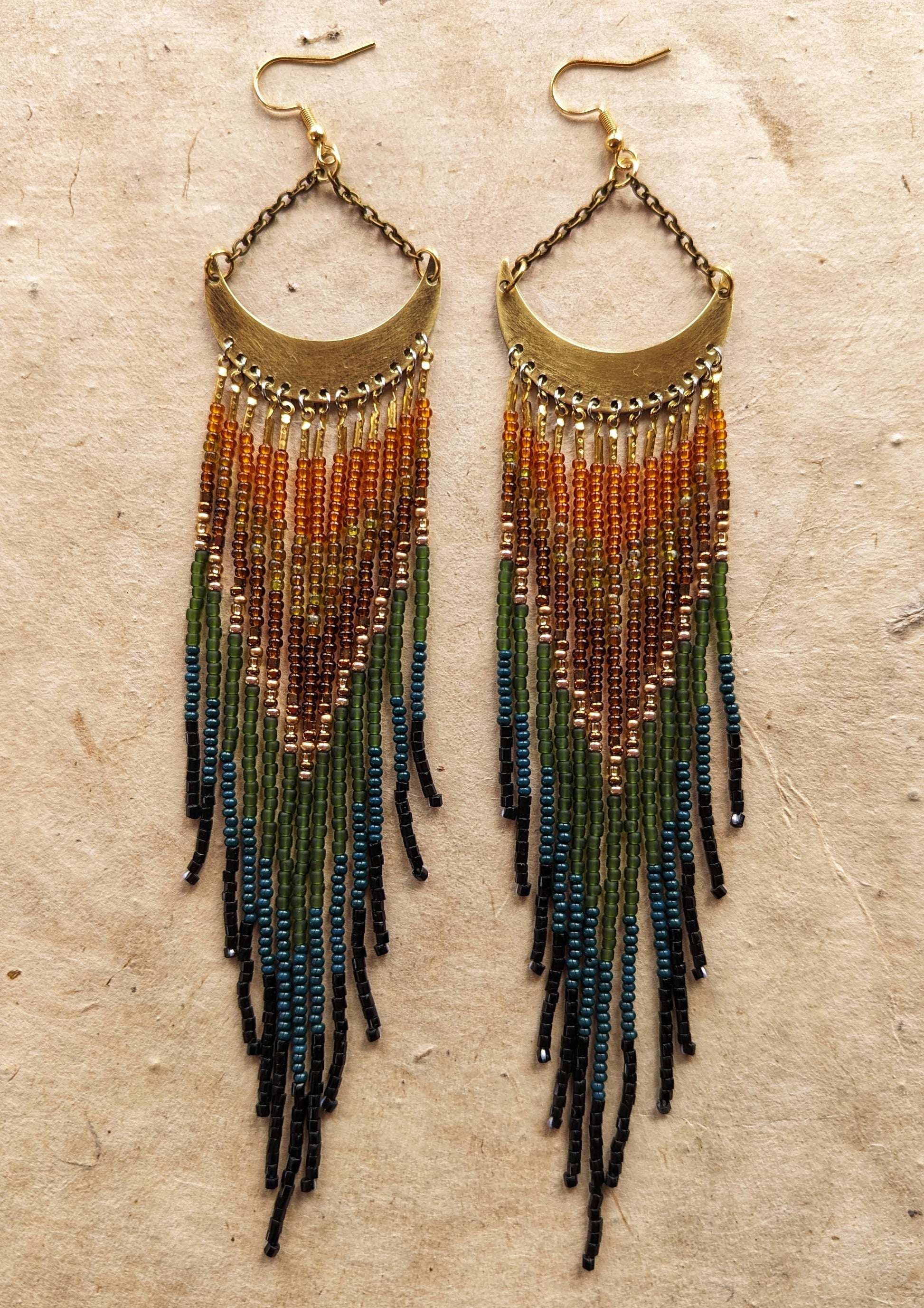 Moon & Milk artisan earrings with brass crescent tops and flowing fringes in burnt orange, amber, olive, dark green, and black seed beads, displayed on neutral paper.