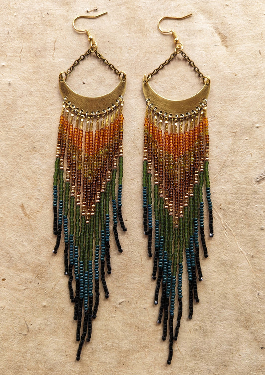 Moon & Milk artisan earrings with brass crescent tops and flowing fringes in burnt orange, amber, olive, dark green, and black seed beads, displayed on neutral paper.