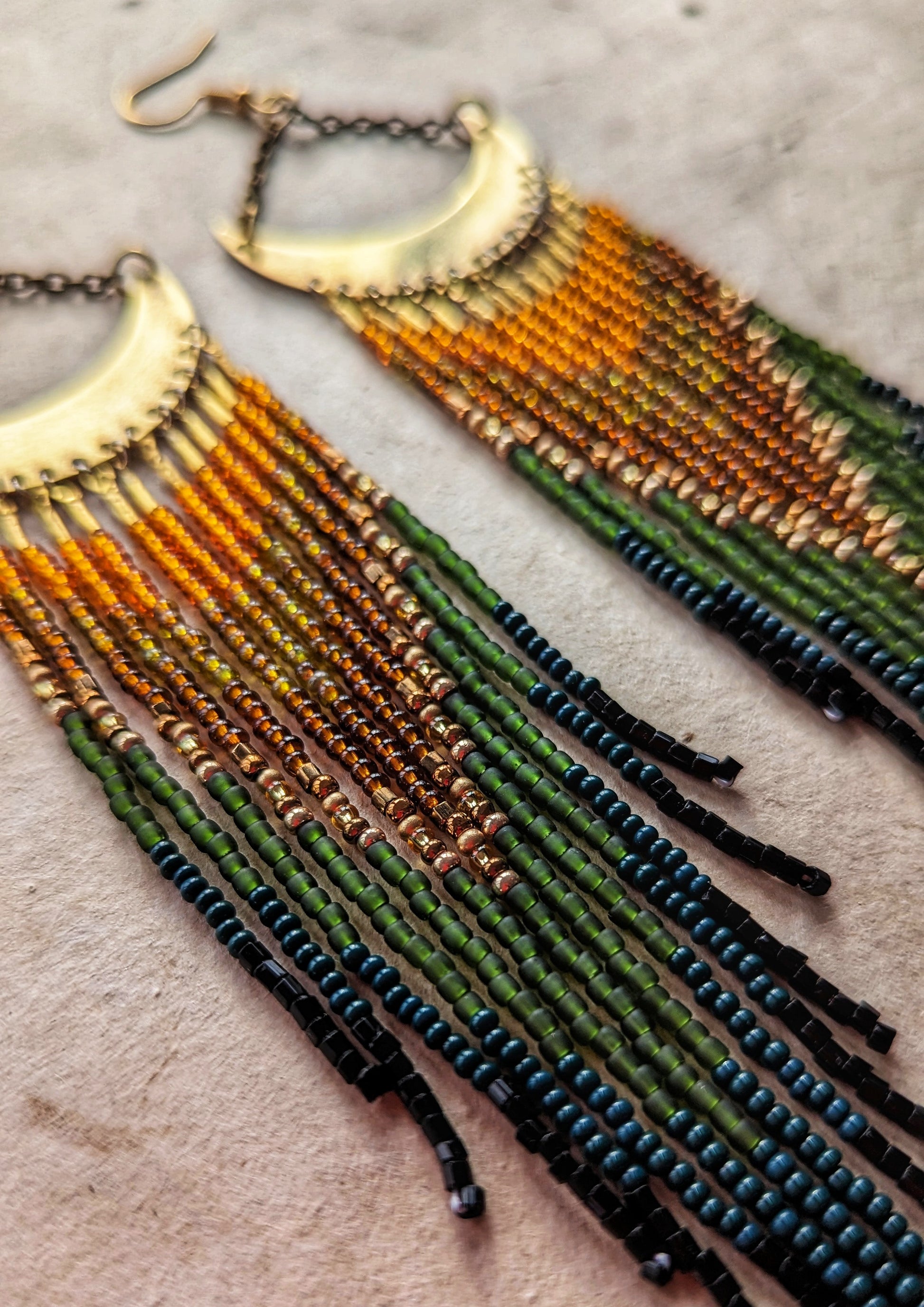 Moon & Milk artisan earrings with brass crescent tops and flowing fringes in burnt orange, amber, olive, dark green, and black seed beads, displayed on neutral paper.
