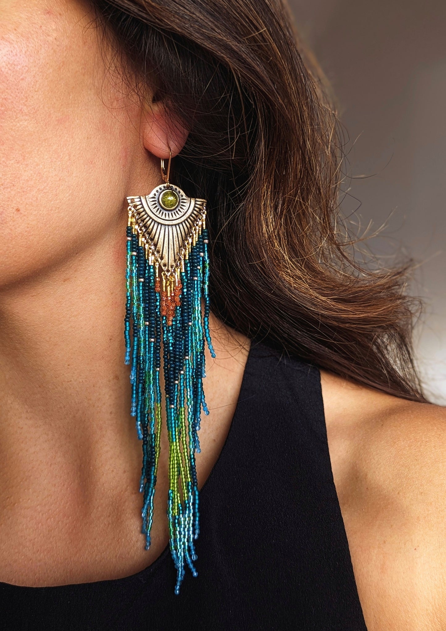 Close-up of long, beaded earrings in blue, green, and rust tones, with a brass sunburst design and a green stone, worn by a person with wavy hair, dressed in a black top.