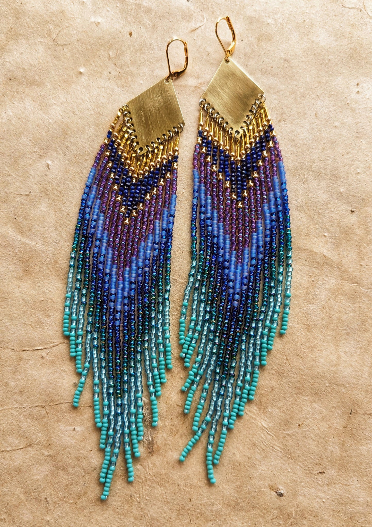 Moon & Milk long beaded earrings in layered blue, turquoise, purple, and brass tones, handmade with brass triangles and inspired by ocean depth and mystery