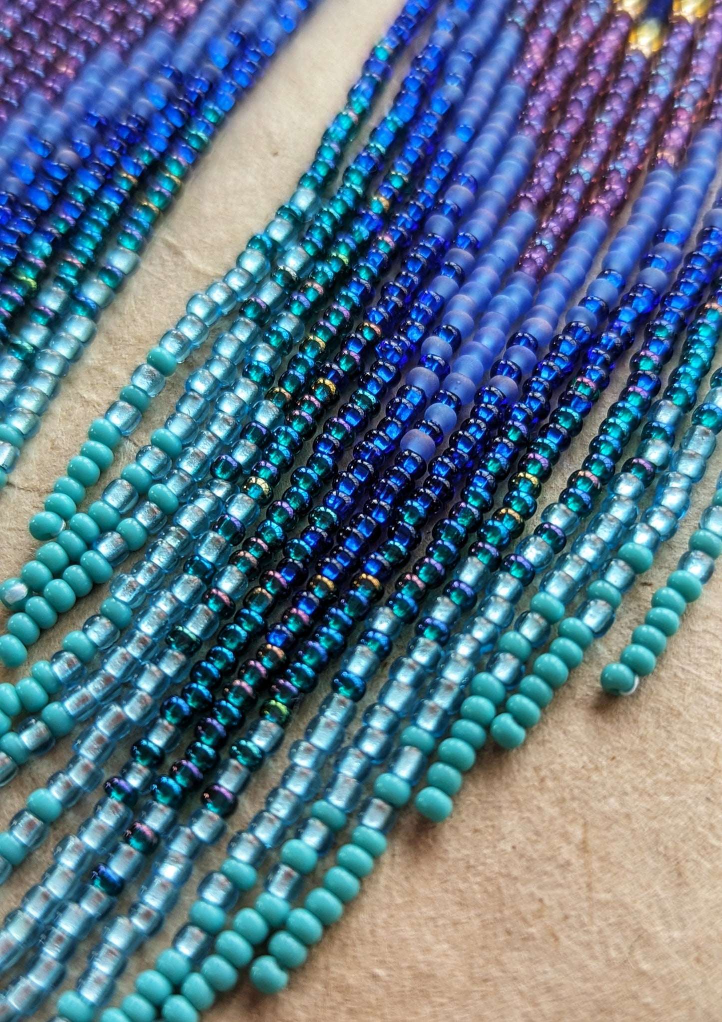 Close-up of long beaded earrings in layered blue, turquoise, purple, and brass tones, handmade with brass triangles and inspired by ocean depth and mystery.