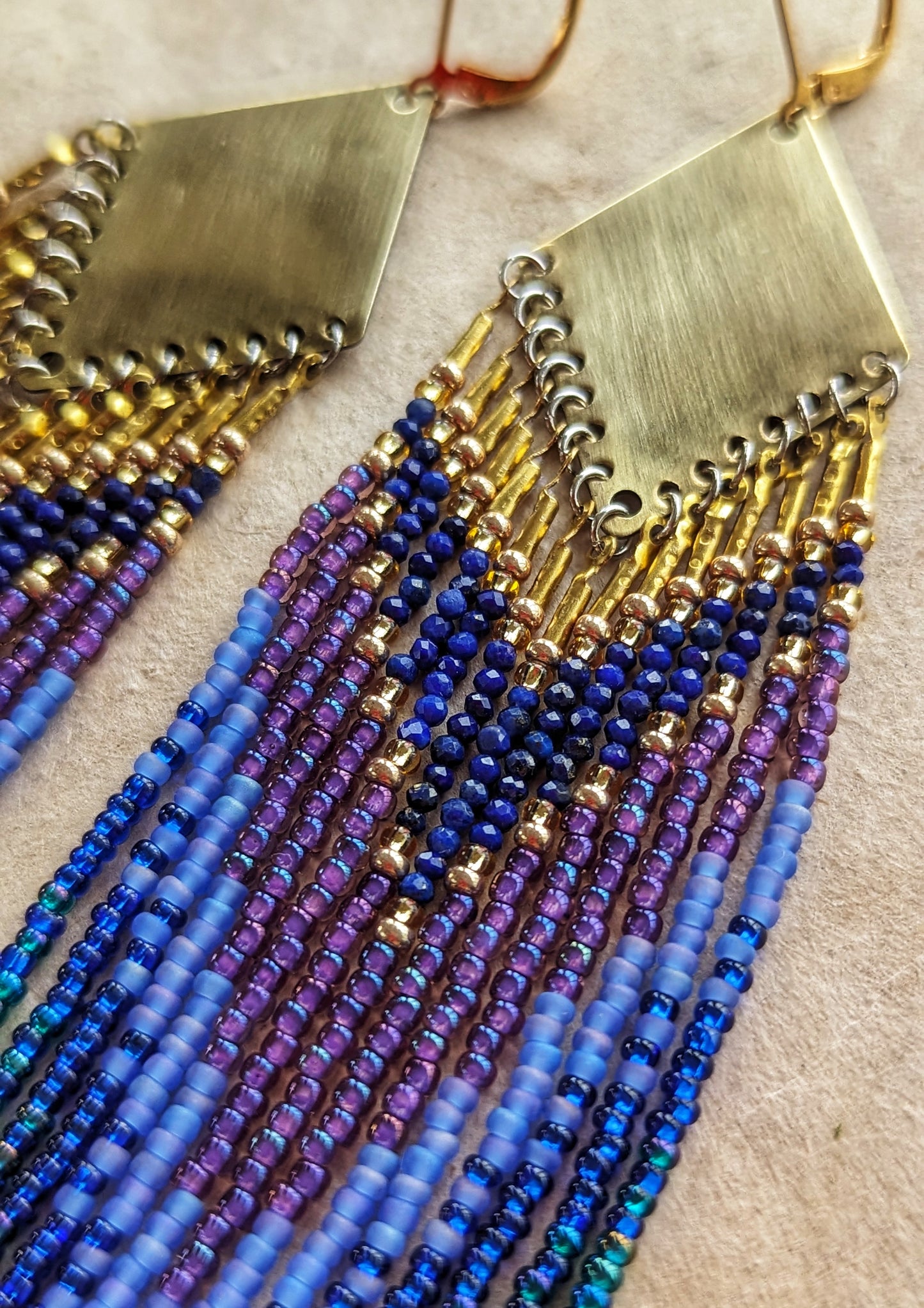 Close-up of long beaded earrings in layered blue, turquoise, purple, and brass tones, handmade with brass triangles and inspired by ocean depth and mystery.