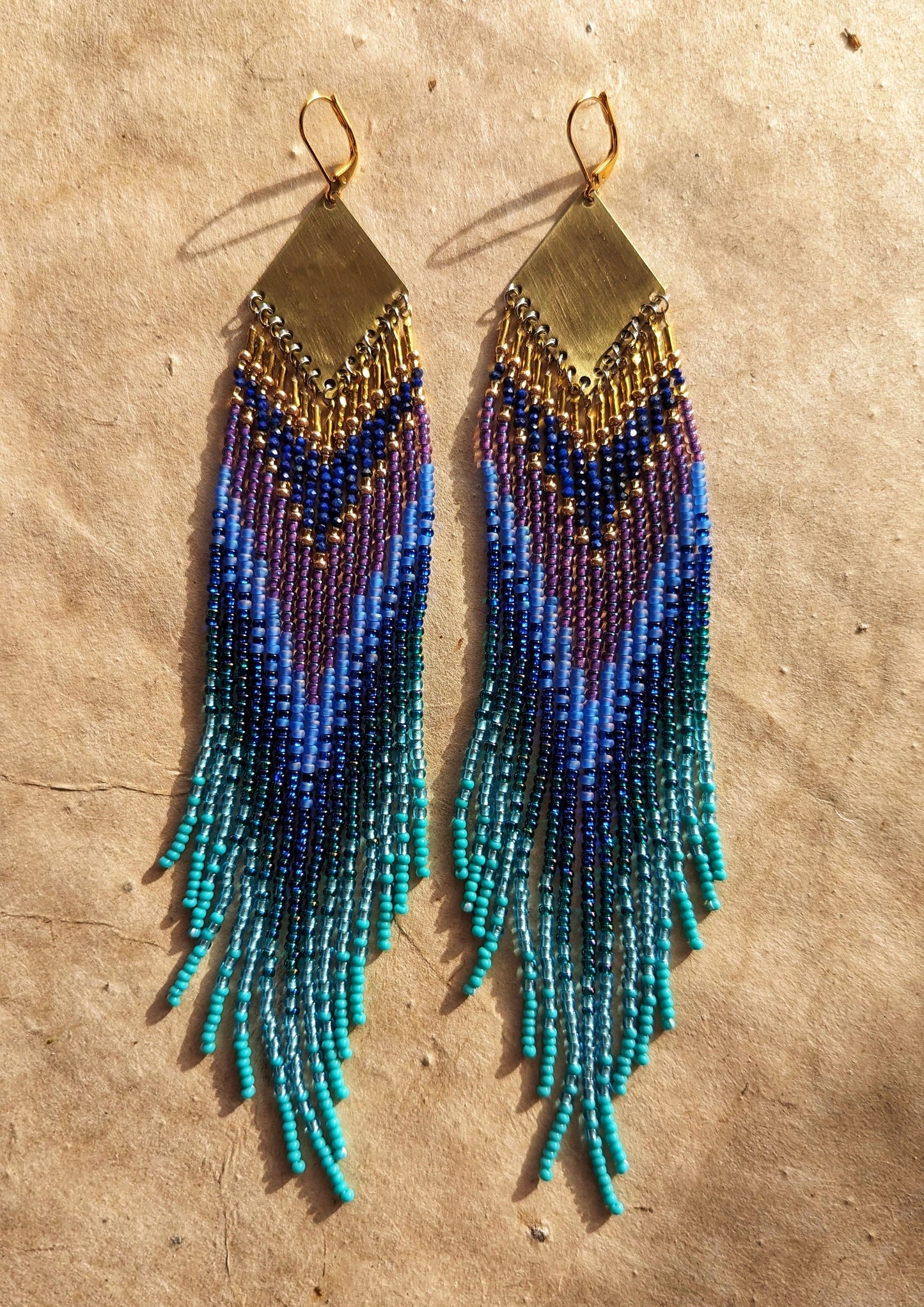 Moon & Milk long beaded earrings in layered blue, turquoise, purple, and brass tones, handmade with brass triangles and inspired by ocean depth and mystery