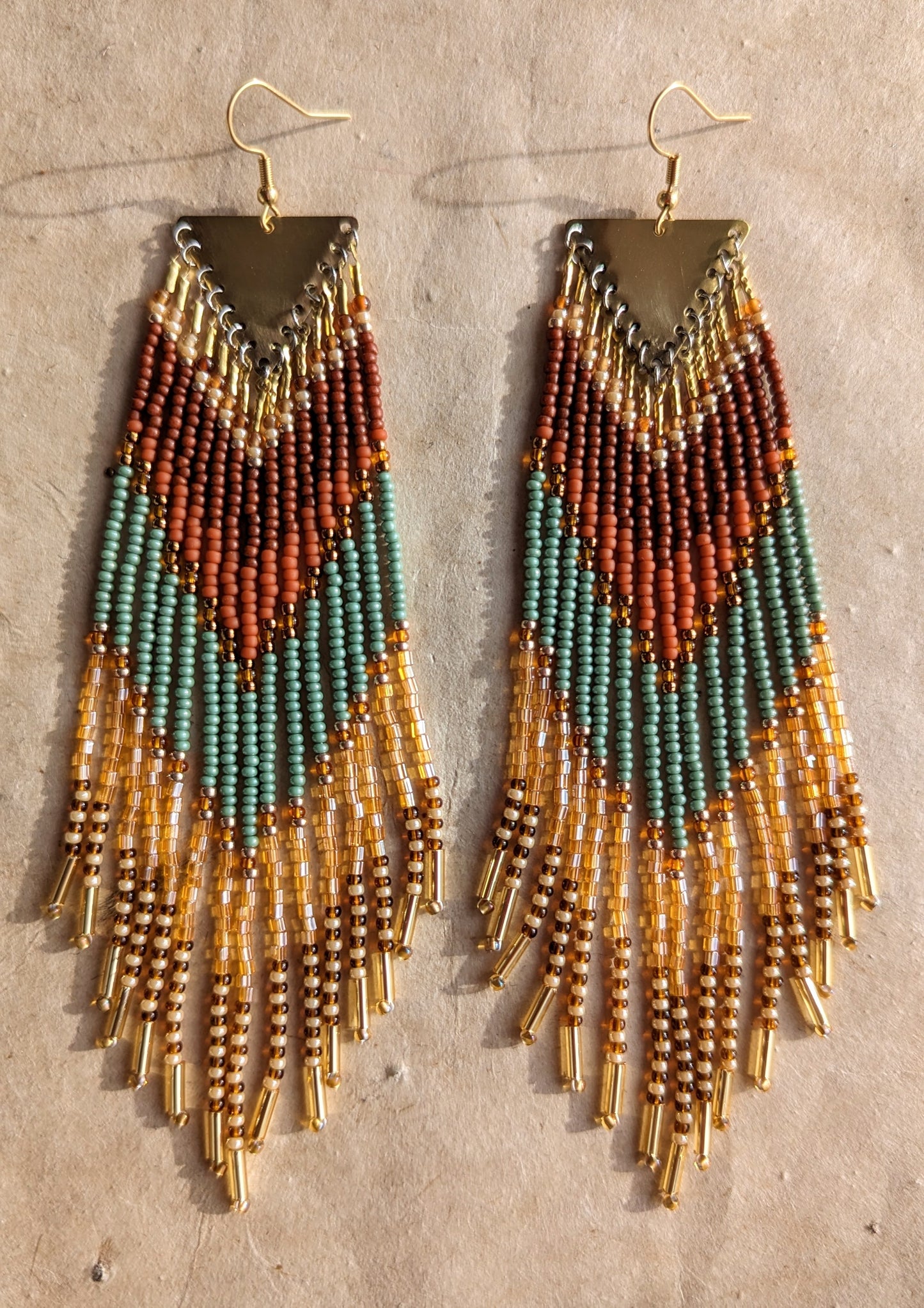 Pair of beaded earrings with a triangular brass top, featuring earthy tones of terracotta, green, and amber with dangling gold tube beads at the ends, placed on a beige background. - Moon & Milk