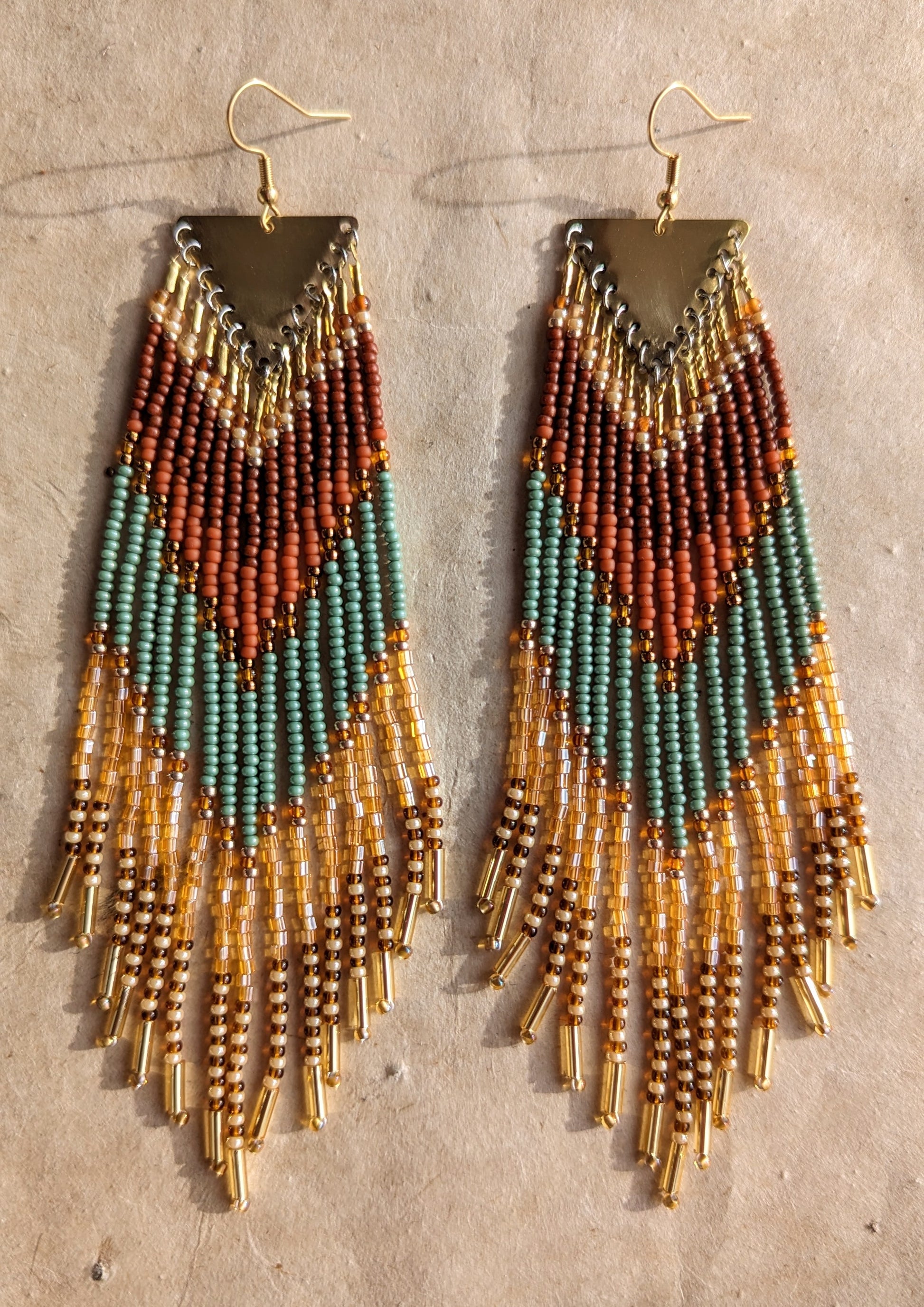 Pair of beaded earrings with a triangular brass top, featuring earthy tones of terracotta, green, and amber with dangling gold tube beads at the ends, placed on a beige background. - Moon & Milk