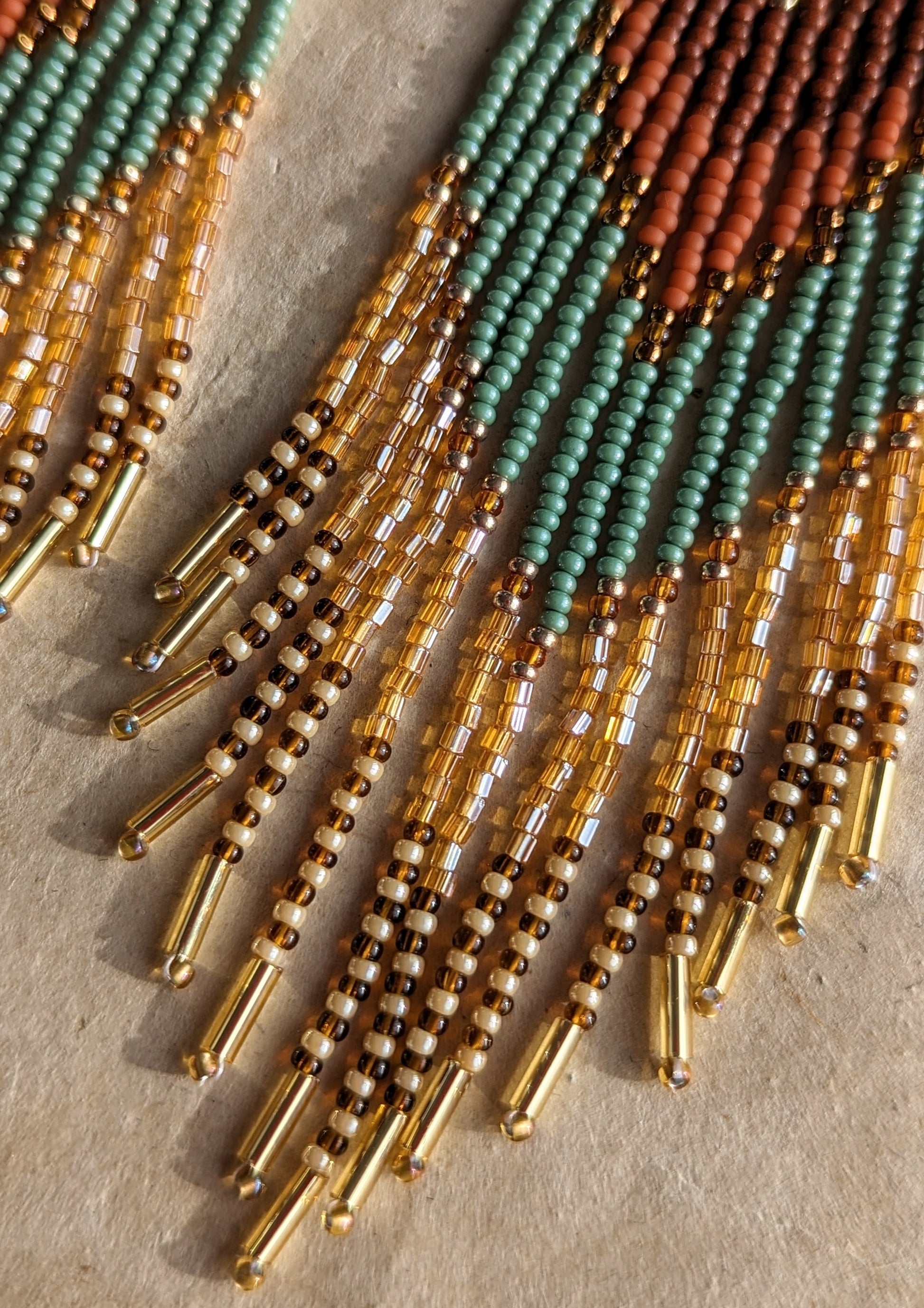 Close-up of the beaded earrings showing the intricate details of terracotta, green, and amber beads with gold accents and gold tube beads at the ends on a beige background. - Moon & Milk