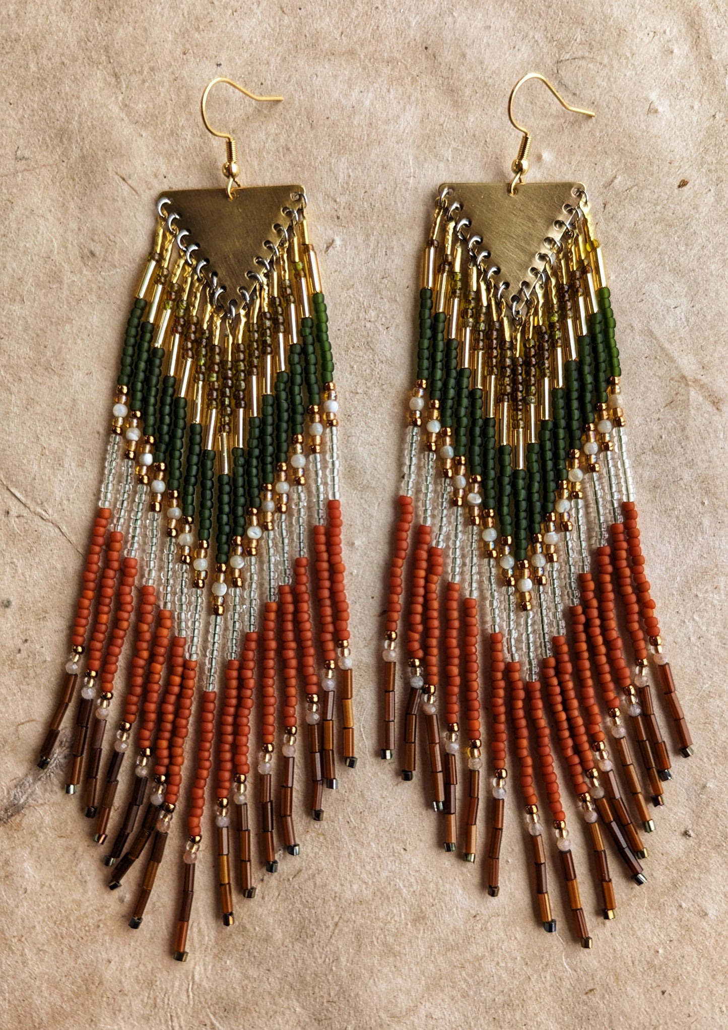 Moon & Milk - Long seed beaded earrings with green, gold, transparent, and terracotta beads arranged in a chevron pattern. Gold triangular brass charms. Photographed on a beige background.