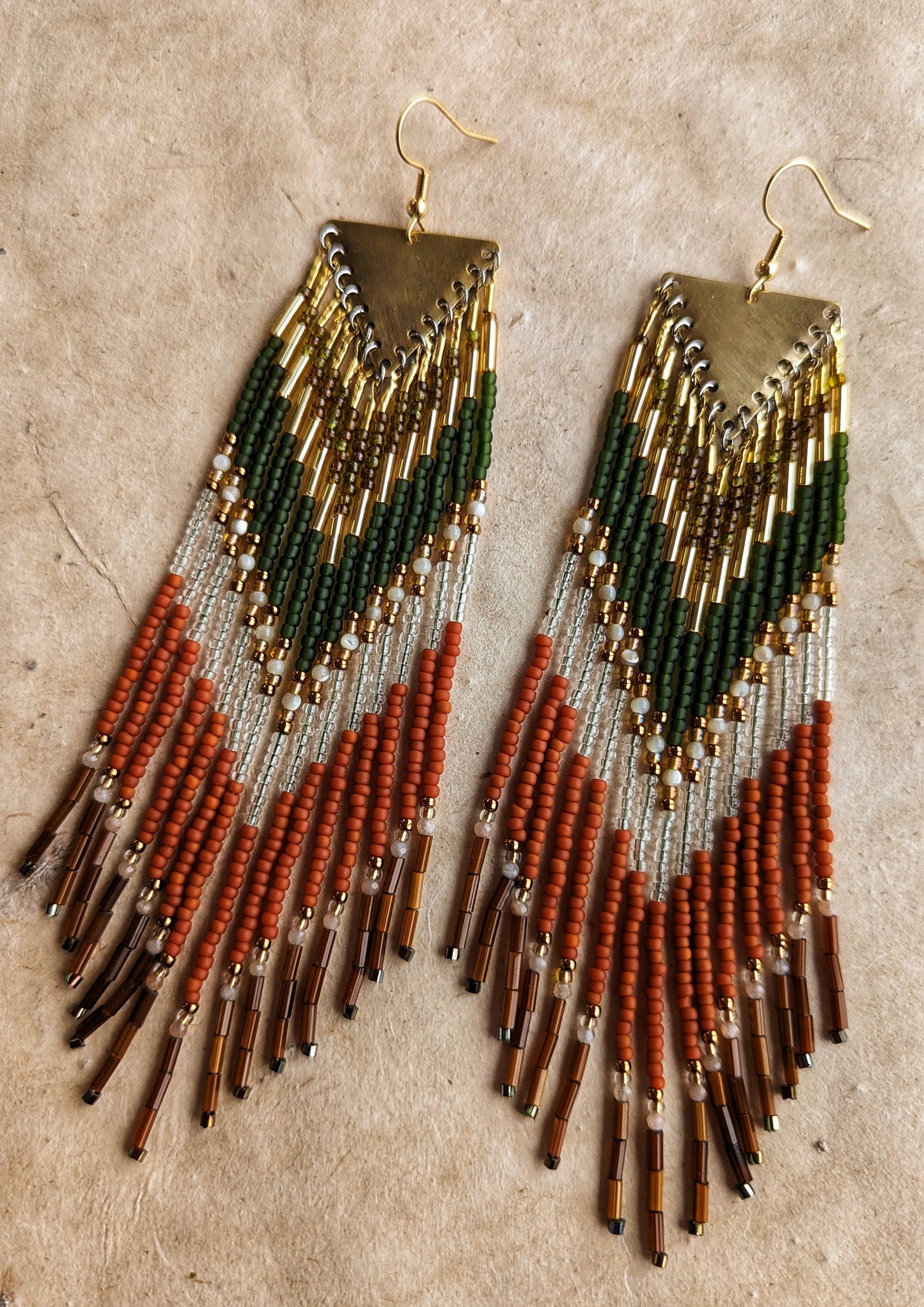 Moon & Milk - Long seed beaded earrings with green, gold, transparent, and terracotta beads arranged in a chevron pattern. Gold triangular brass charms. Photographed on a beige background.