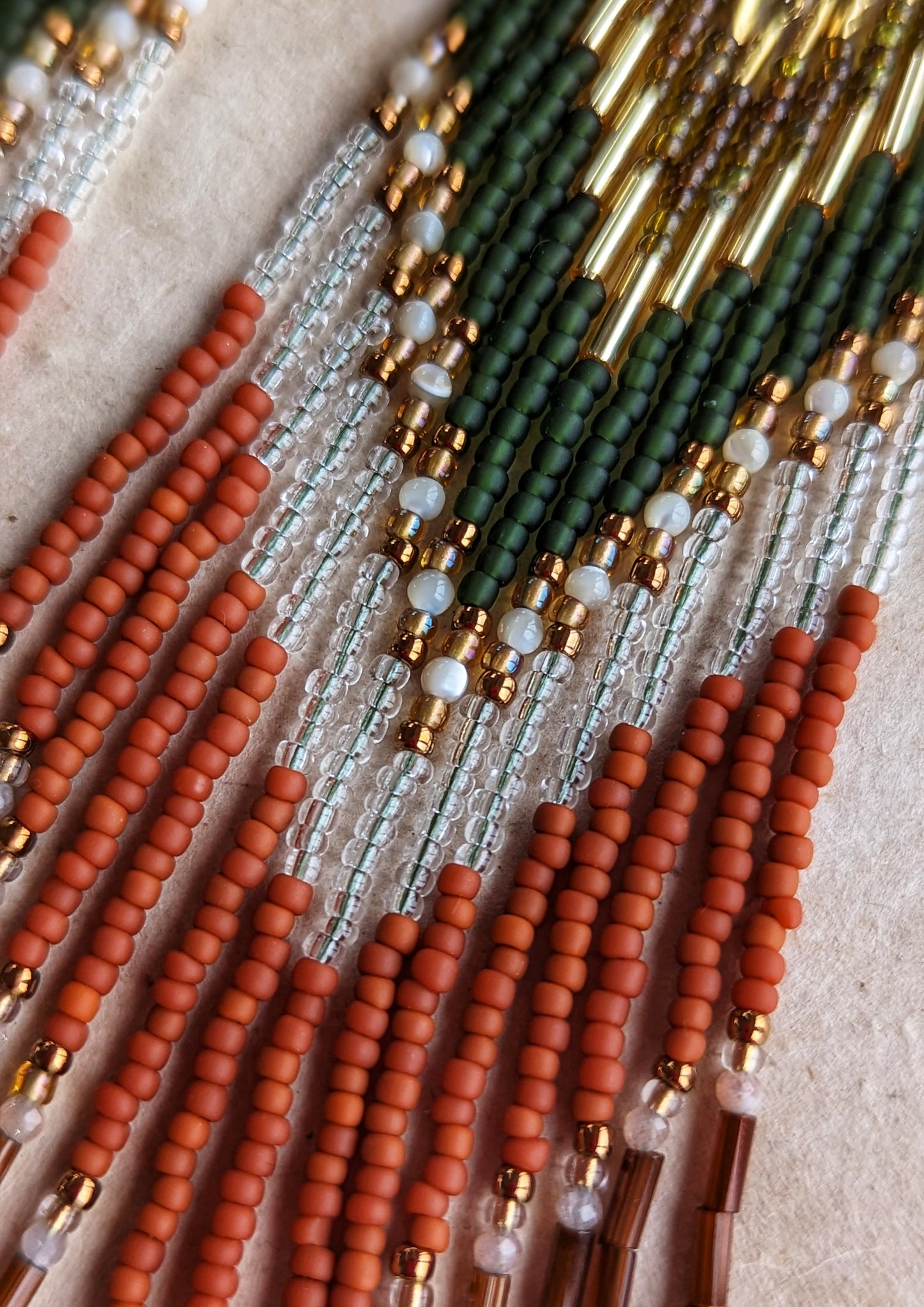 Moon & Milk - Long seed beaded earrings with green, gold, transparent, and terracotta beads arranged in a chevron pattern. Gold triangular brass charms. Photographed on a beige background.