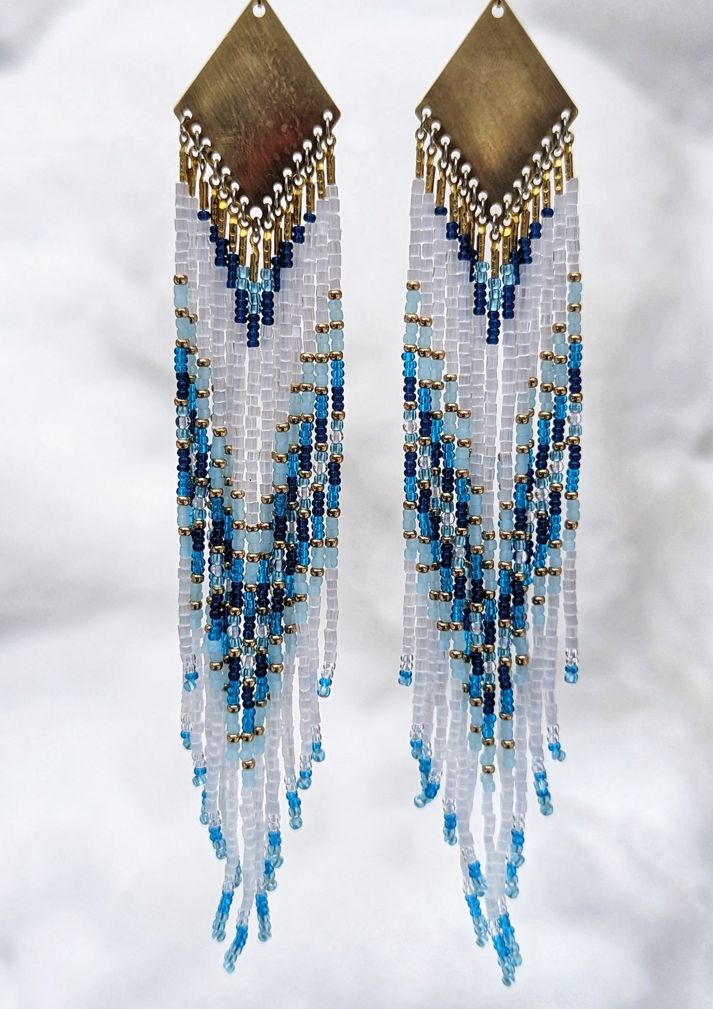 Seed bead earrings with white and blue beaded strands in a gradient pattern with a diamond-shaped golden brass charm.