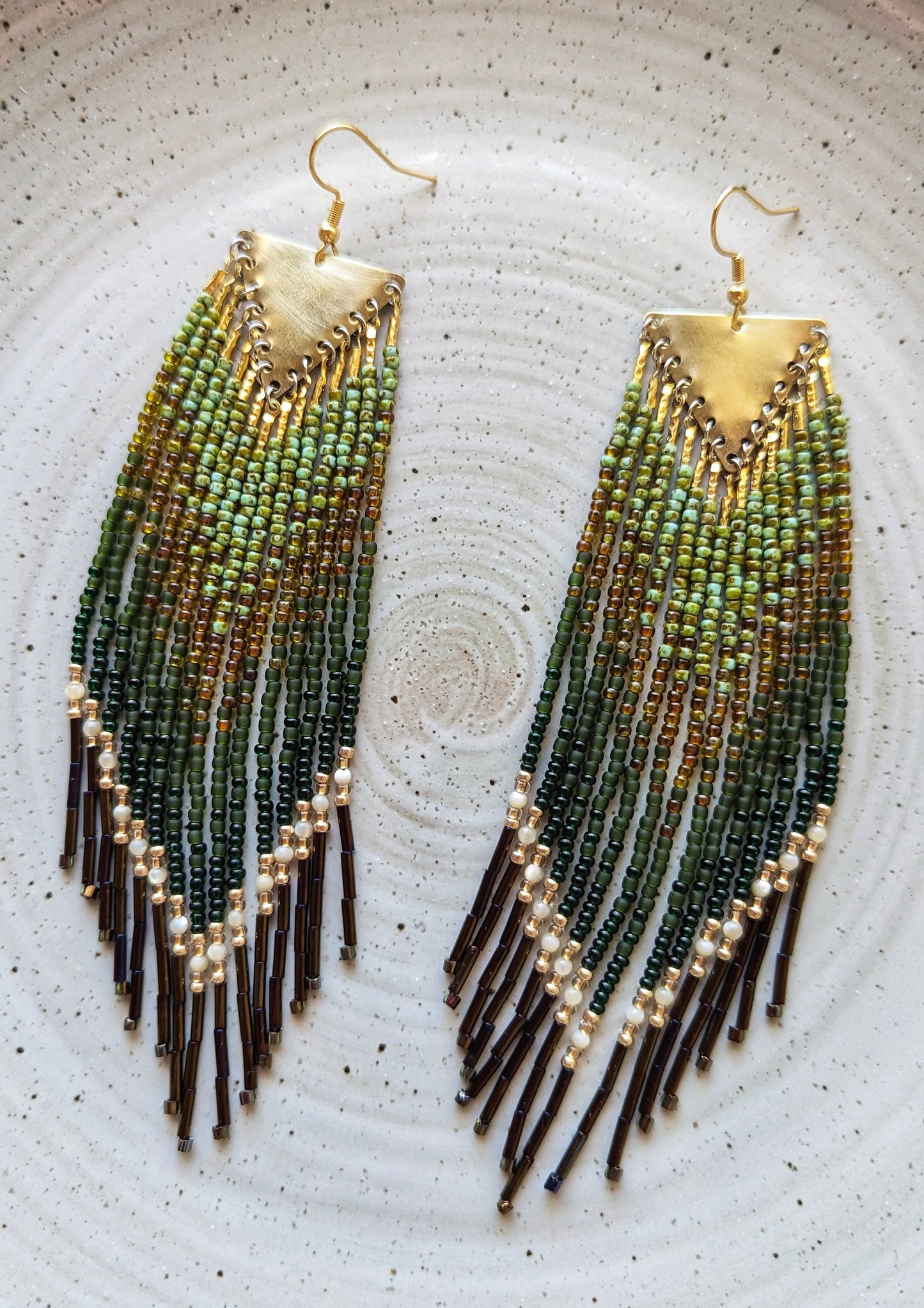 Long native beaded earrings by Moon & Milk in forest green, olive, and gold with pearl accents and black bugle tips, placed on a ceramic plate.