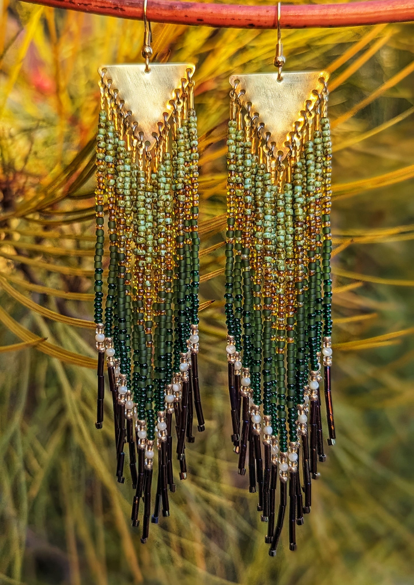 Long native beaded earrings in forest green, olive, and gold with pearl accents and bronze bugle tips, hanging against soft golden foliage.