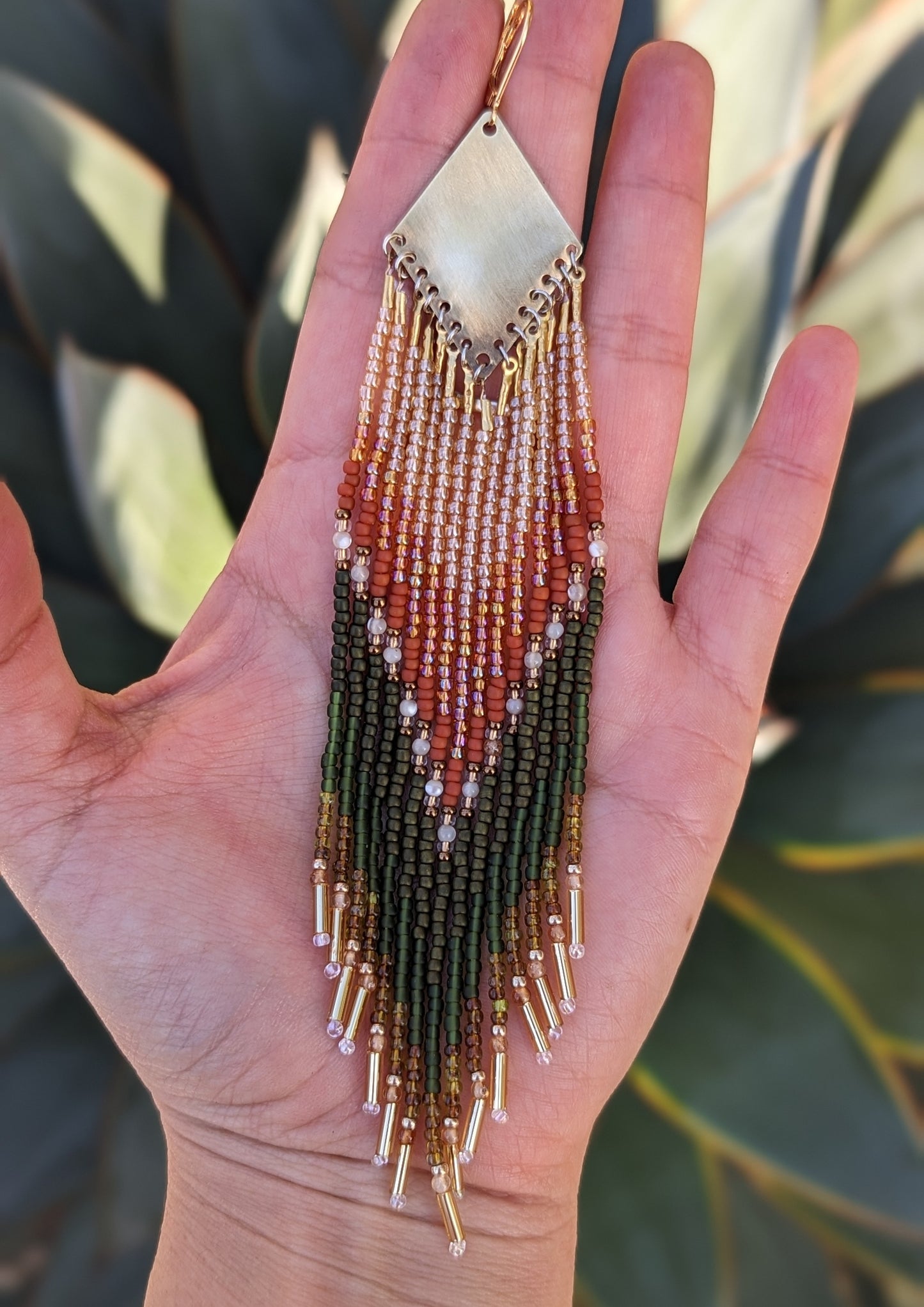 A single, handcrafted earring held in the palm of a hand, showcasing intricate beadwork in earthy tones of green, orange, and brown, with hints of gold. The beads form a beautiful chevron pattern that flows into dangling fringes, giving the earring an elegant, nature-inspired look. - Moon & Milk