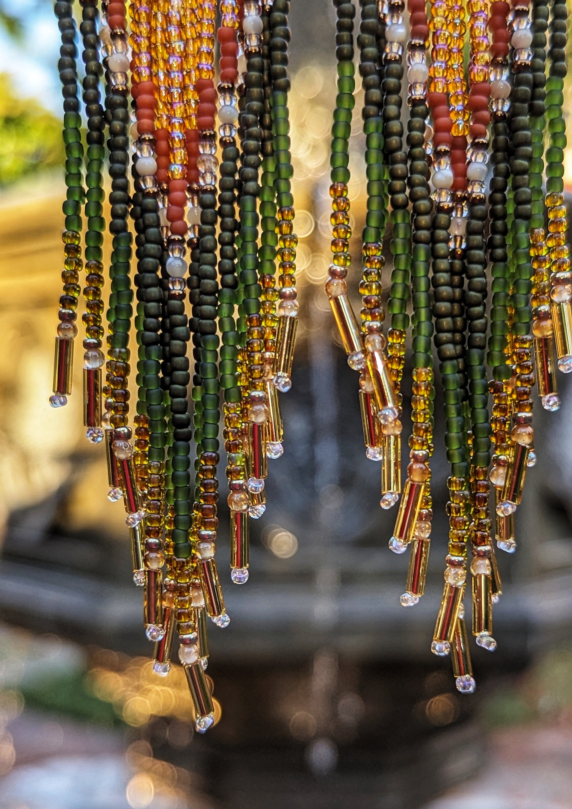 A close-up of the earring fringes against a blurred background, highlighting the vibrant beads in green, orange, and gold. The beads catch the light, creating a sparkling effect that emphasizes the fine detail and craftsmanship of each strand. - Moon & Milk