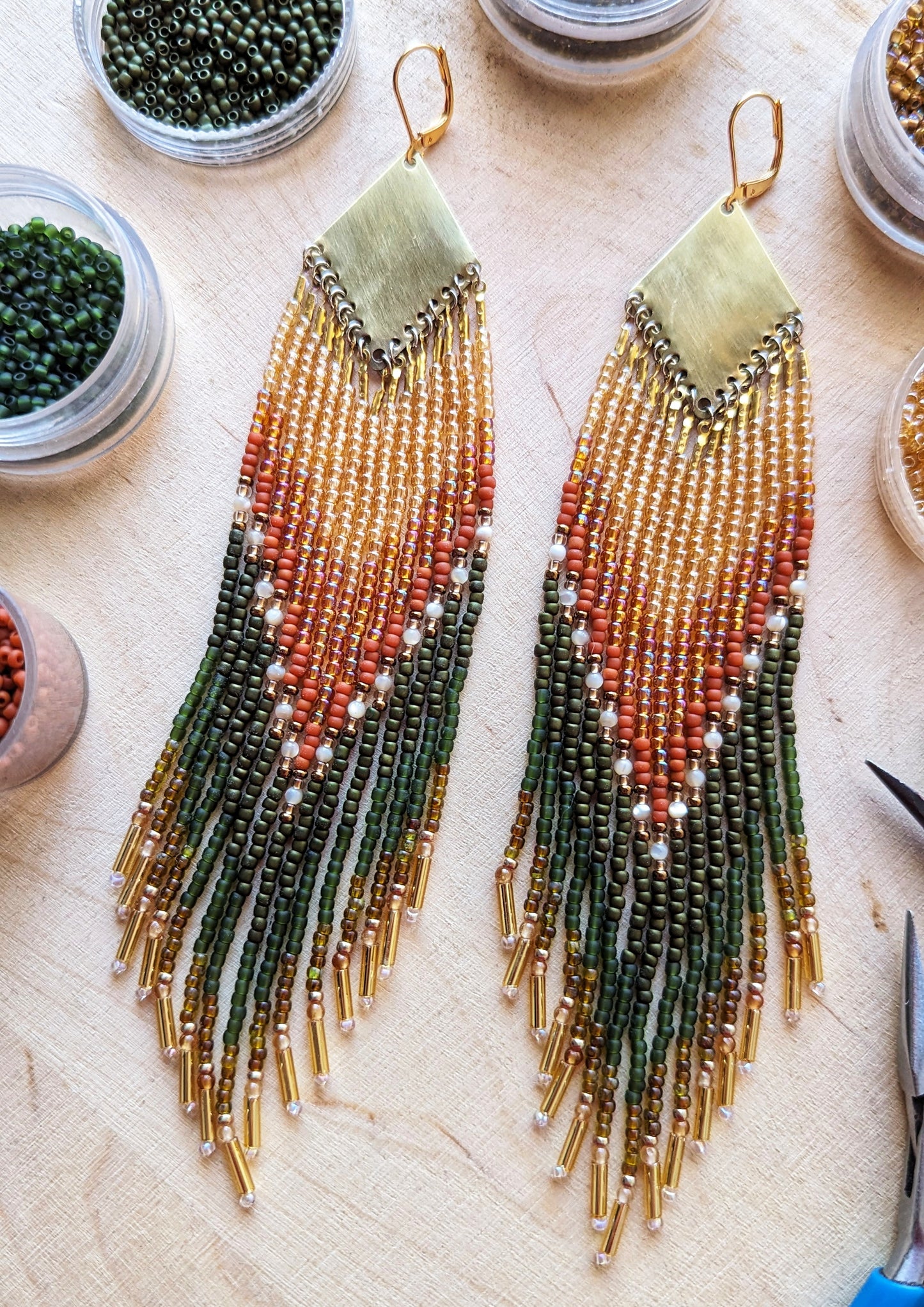 A pair of beaded earrings lying on a light wooden surface, surrounded by small containers of beads. These earrings feature warm tones of green, orange, and gold, with delicate beadwork that creates a cascading effect down to the fringed ends. - Moon & Milk