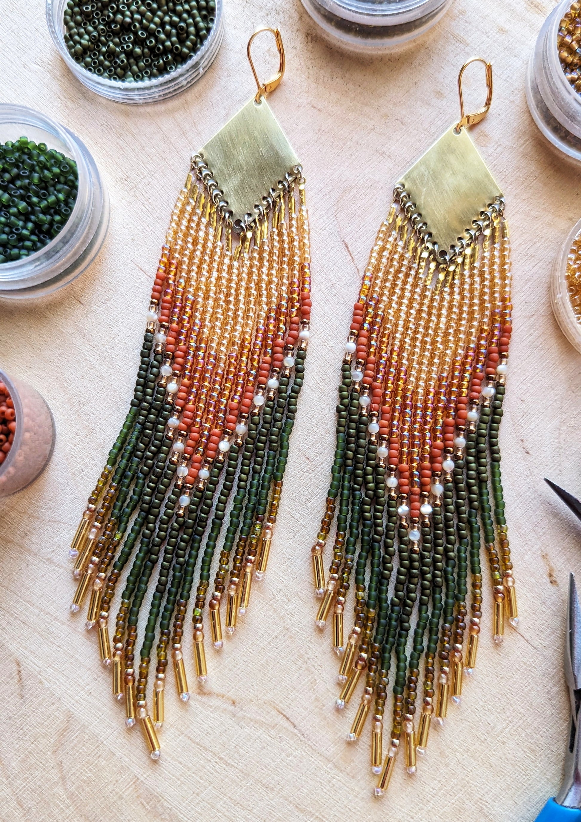 A pair of beaded earrings lying on a light wooden surface, surrounded by small containers of beads. These earrings feature warm tones of green, orange, and gold, with delicate beadwork that creates a cascading effect down to the fringed ends. - Moon & Milk