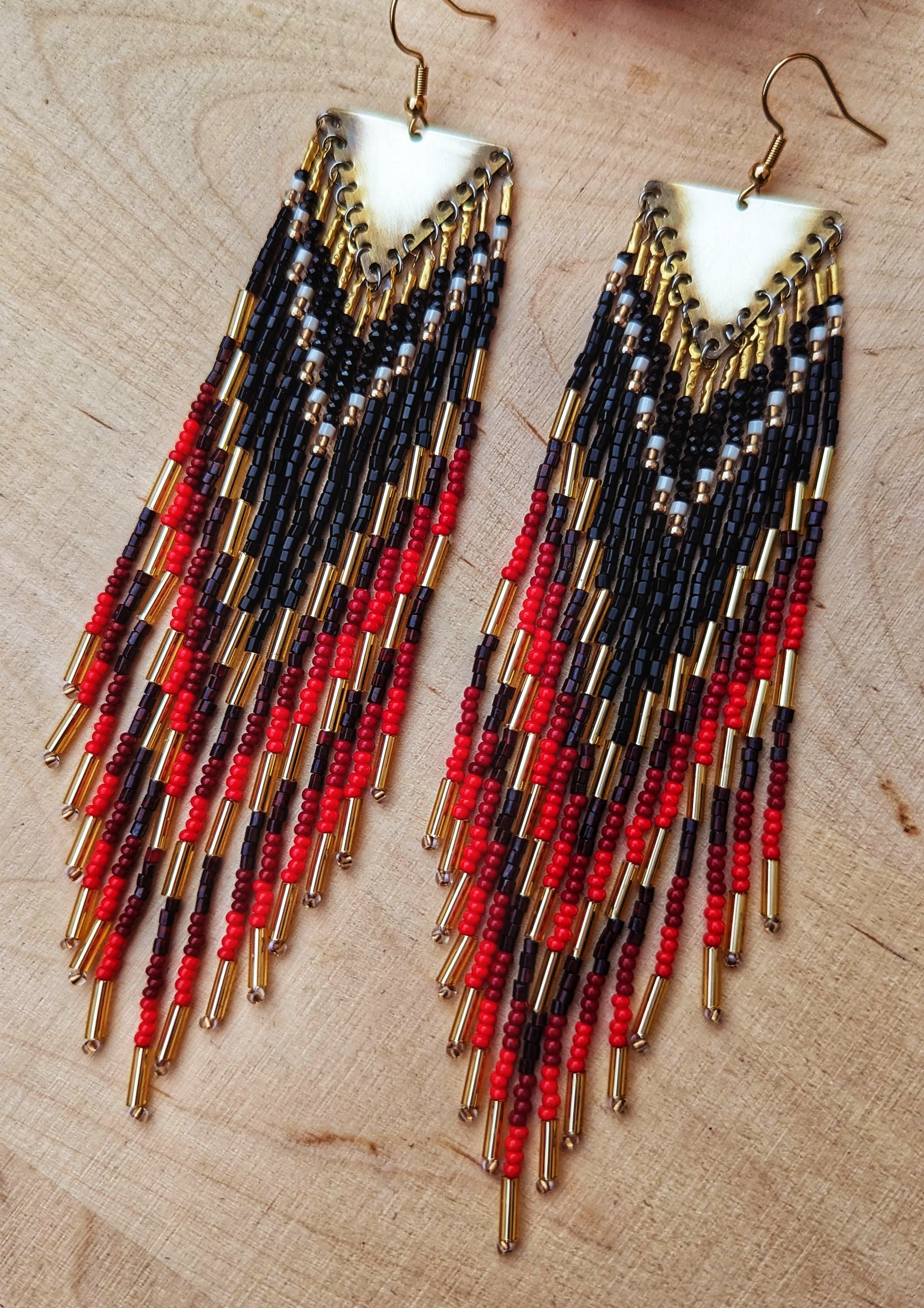 Moon & Milk earrings featuring black, red, and gold seed beads with brass triangle tops, laid on textured earth-toned fabric next to dried grasses.