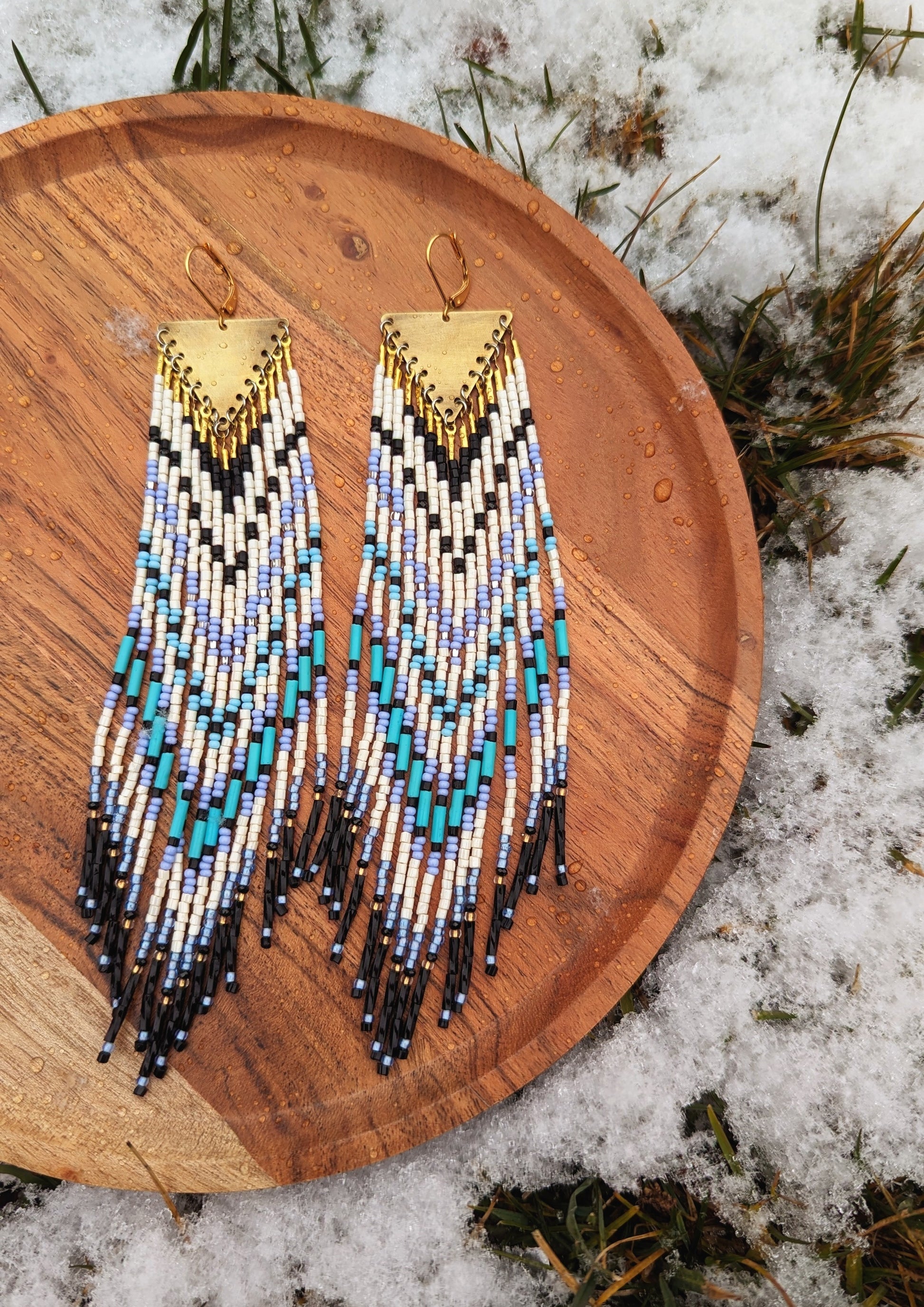 Moon & Milk long native beaded earrings with white, black, icy blue, and turquoise beadwork on brass triangles, displayed on wood with snow.