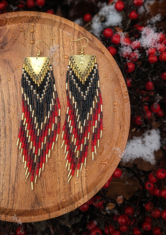 Moon & Milk beaded earrings with black, red, and gold beads, hanging from brass triangles, displayed on a wet wooden surface with berries and snow.