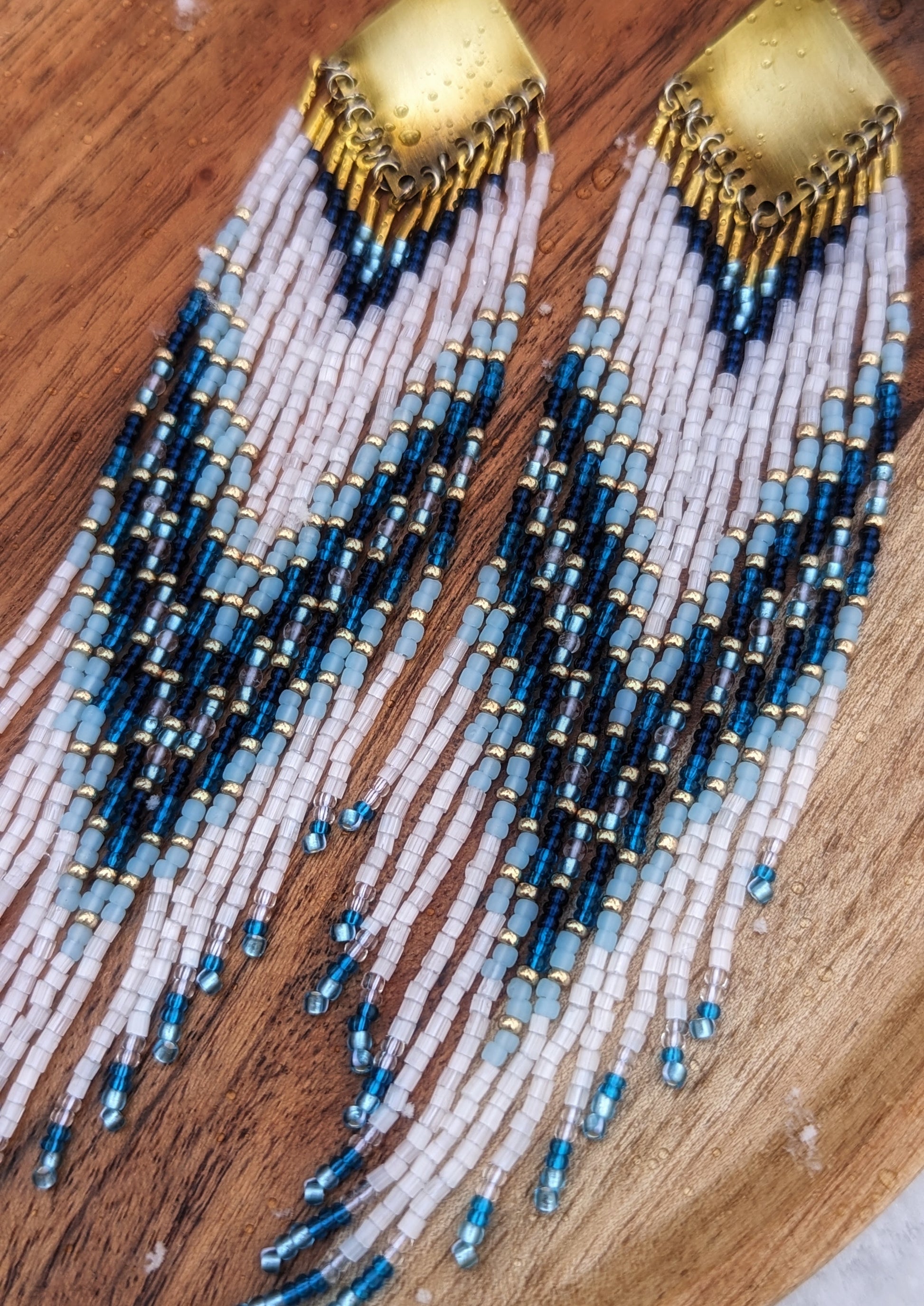 Seed bead earrings with white and blue beaded strands in a gradient pattern with a diamond-shaped golden brass  charm.