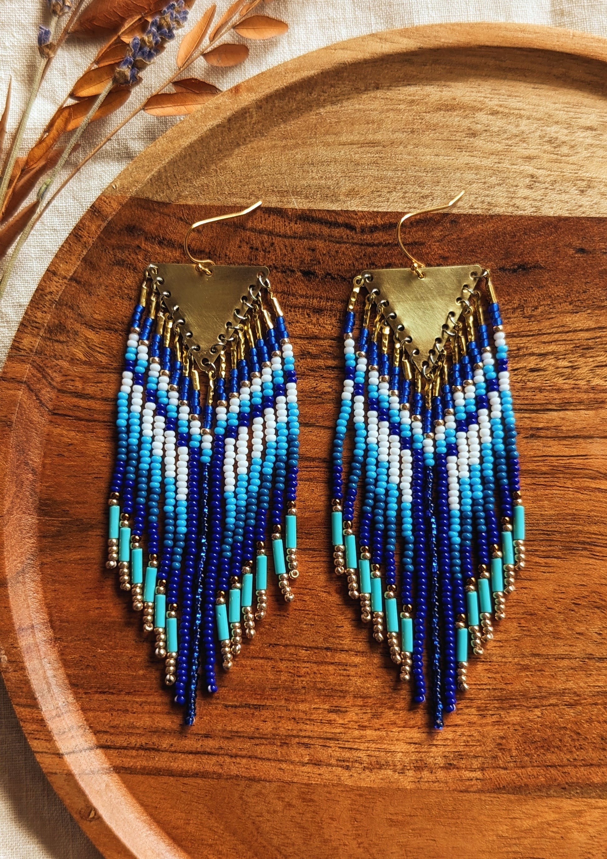 Handcrafted native earrings with cobalt, turquoise, and white beadwork, brass triangle tops, and gold accents. Displayed on a wooden tray with dried lavender.