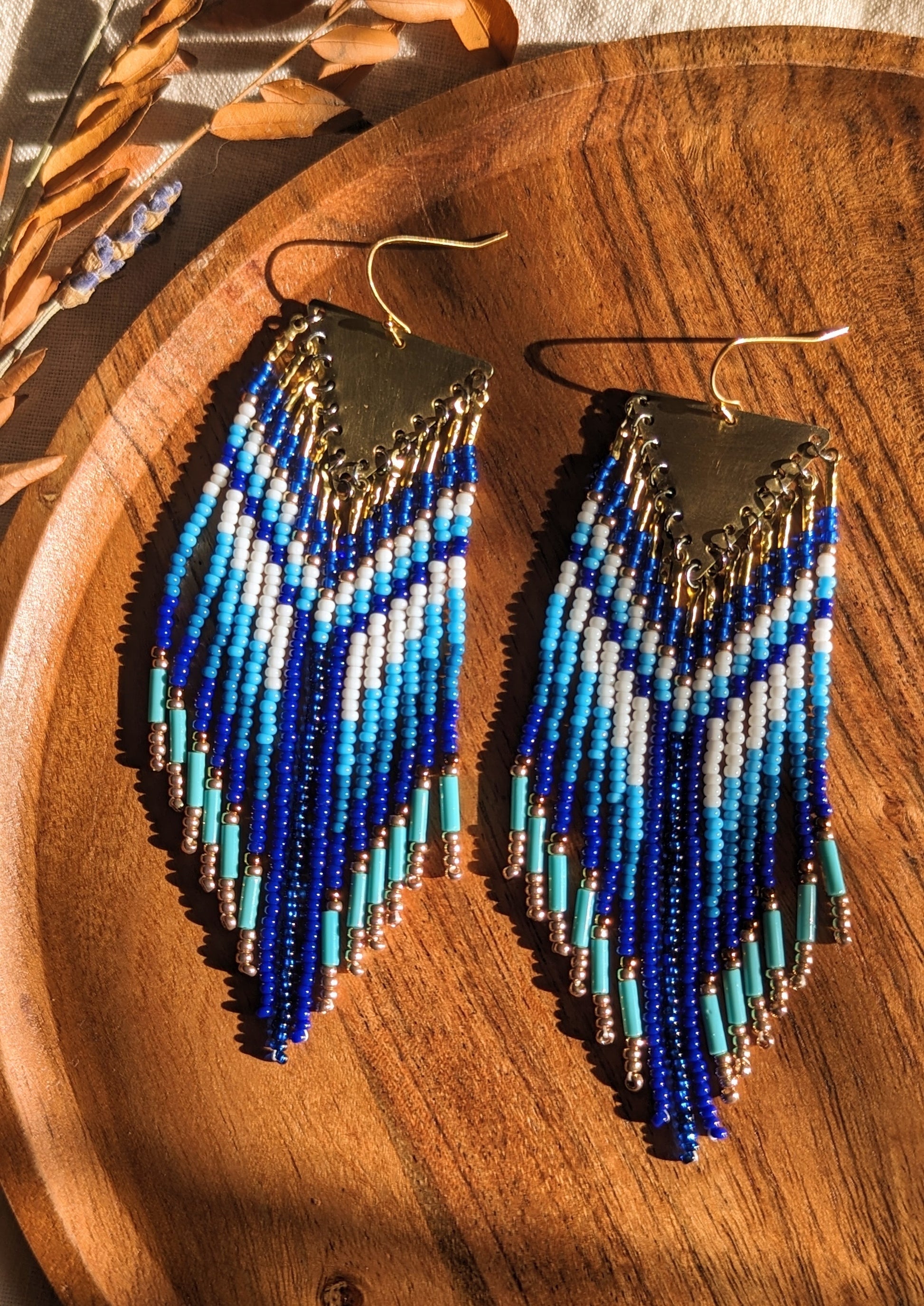 Handcrafted native earrings with cobalt, turquoise, and white beadwork, brass triangle tops, and gold accents. Displayed on a wooden tray with dried lavender.