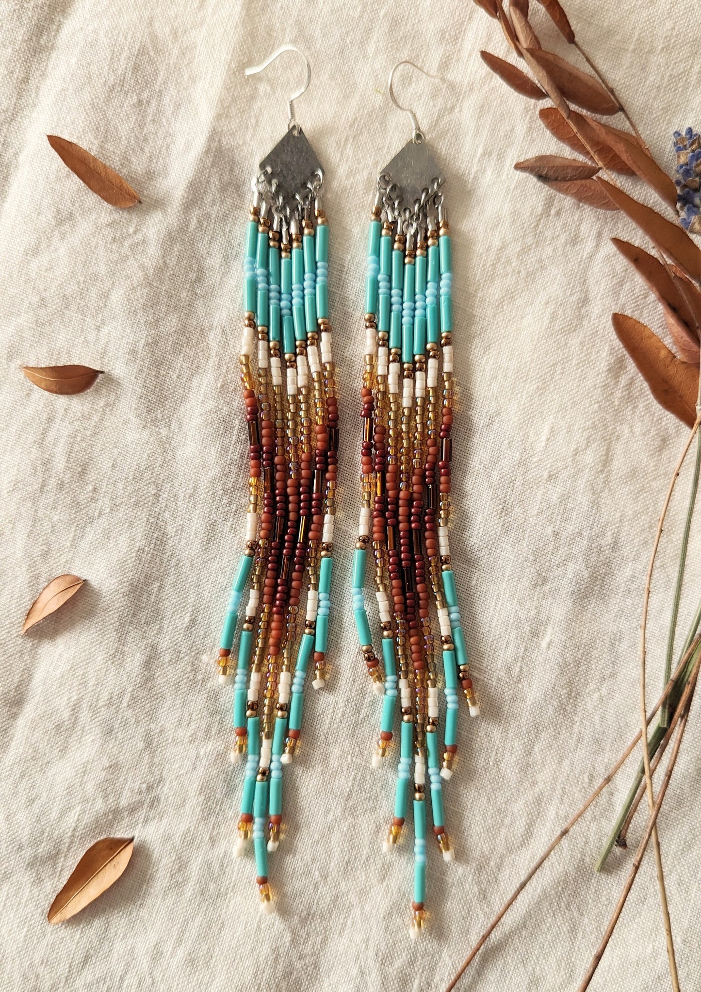 Long beaded earrings with turquoise, brown, and gold accents displayed on a light fabric background, surrounded by dried leaves and twigs.- Moon & Milk