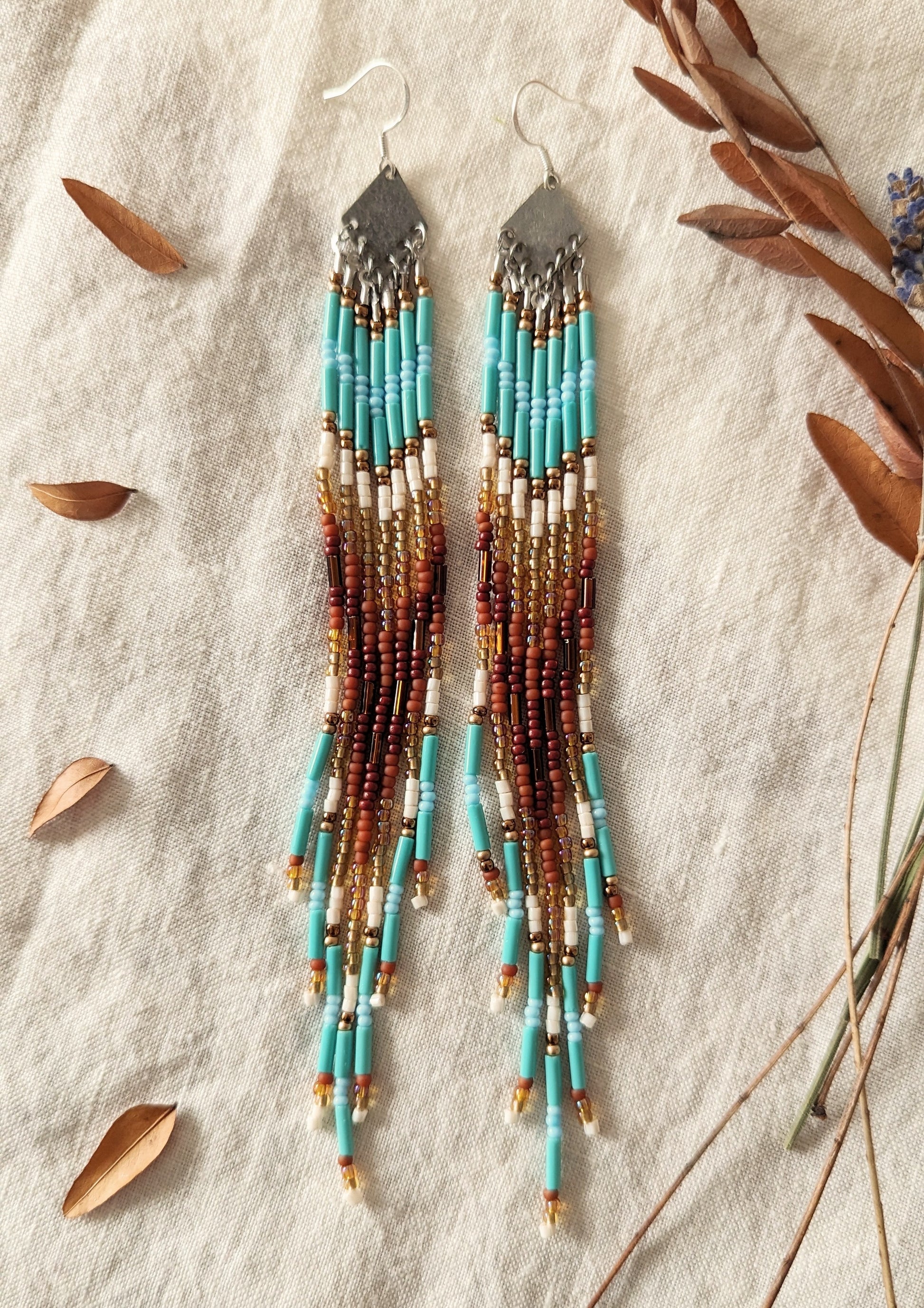Long beaded earrings with turquoise, brown, and gold accents displayed on a light fabric background, surrounded by dried leaves and twigs.- Moon & Milk