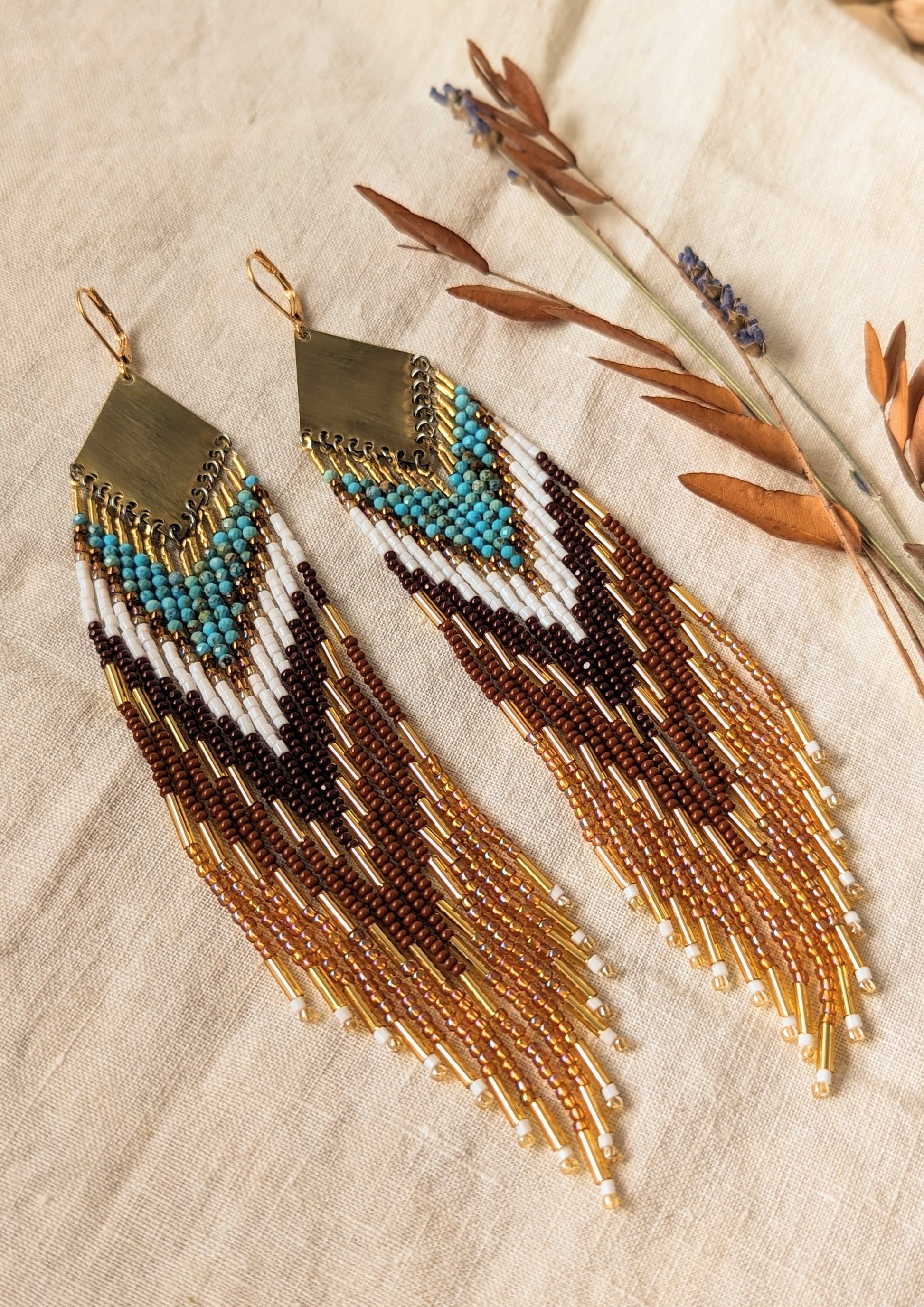 Pair of long beaded earrings with brass tops, featuring shades of turquoise, brown, and gold beads, displayed on a light fabric background with dried leaves on the side. - Moon & Milk