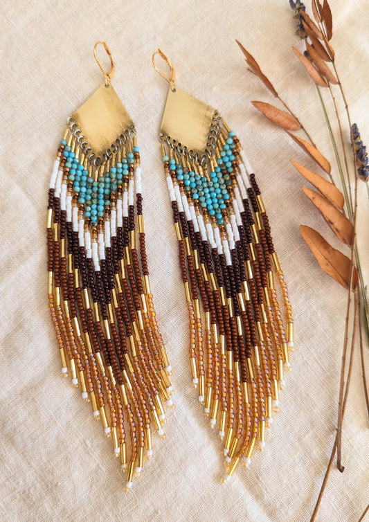 Pair of long beaded earrings with brass tops, featuring shades of turquoise, brown, and gold beads, displayed on a light fabric background with dried leaves on the side. - Moon & Milk