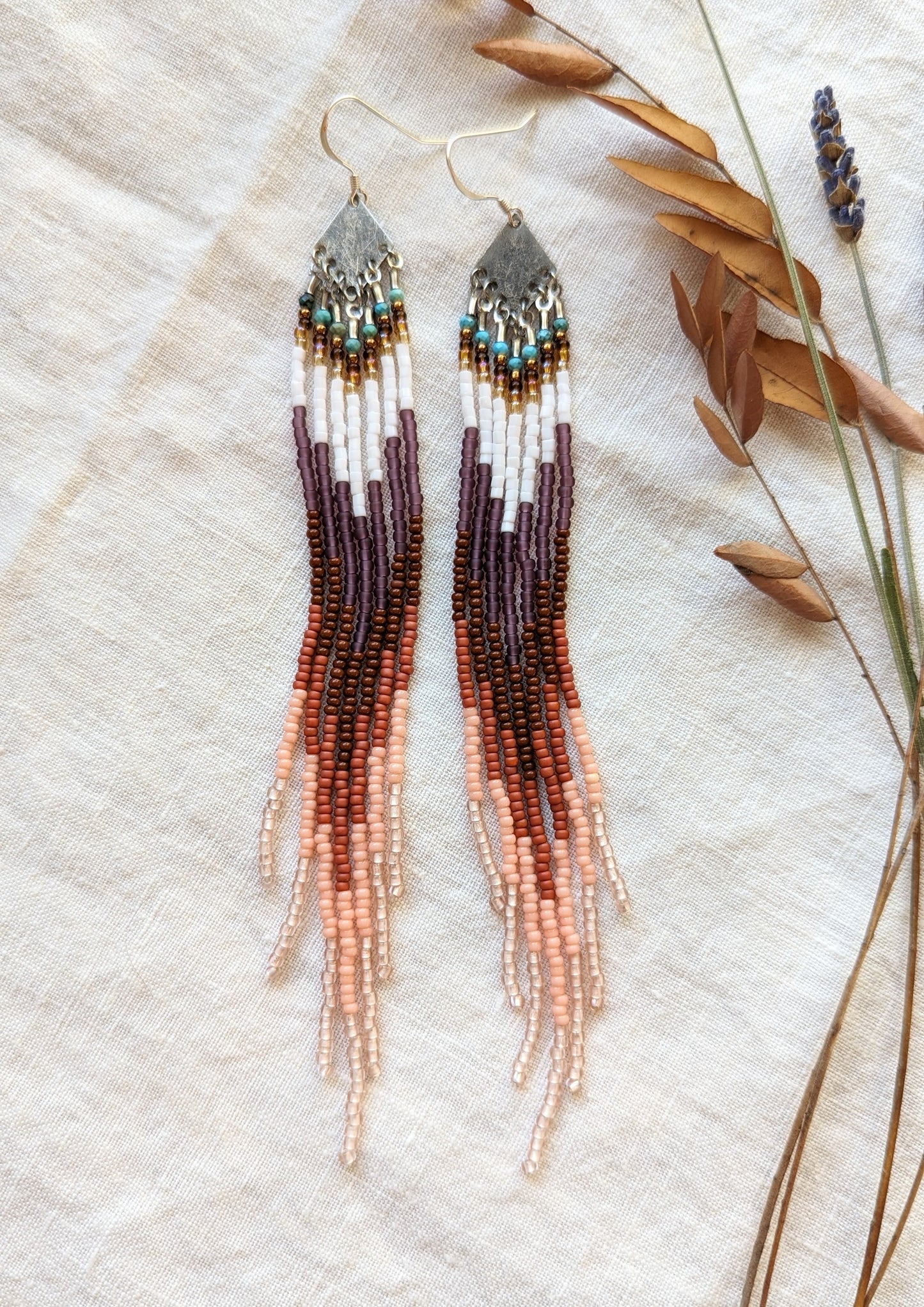 Long beaded fringe earrings with earthy tones of brown, terracotta, and white, displayed on a linen cloth with dried floral accents.-Moon & Milk