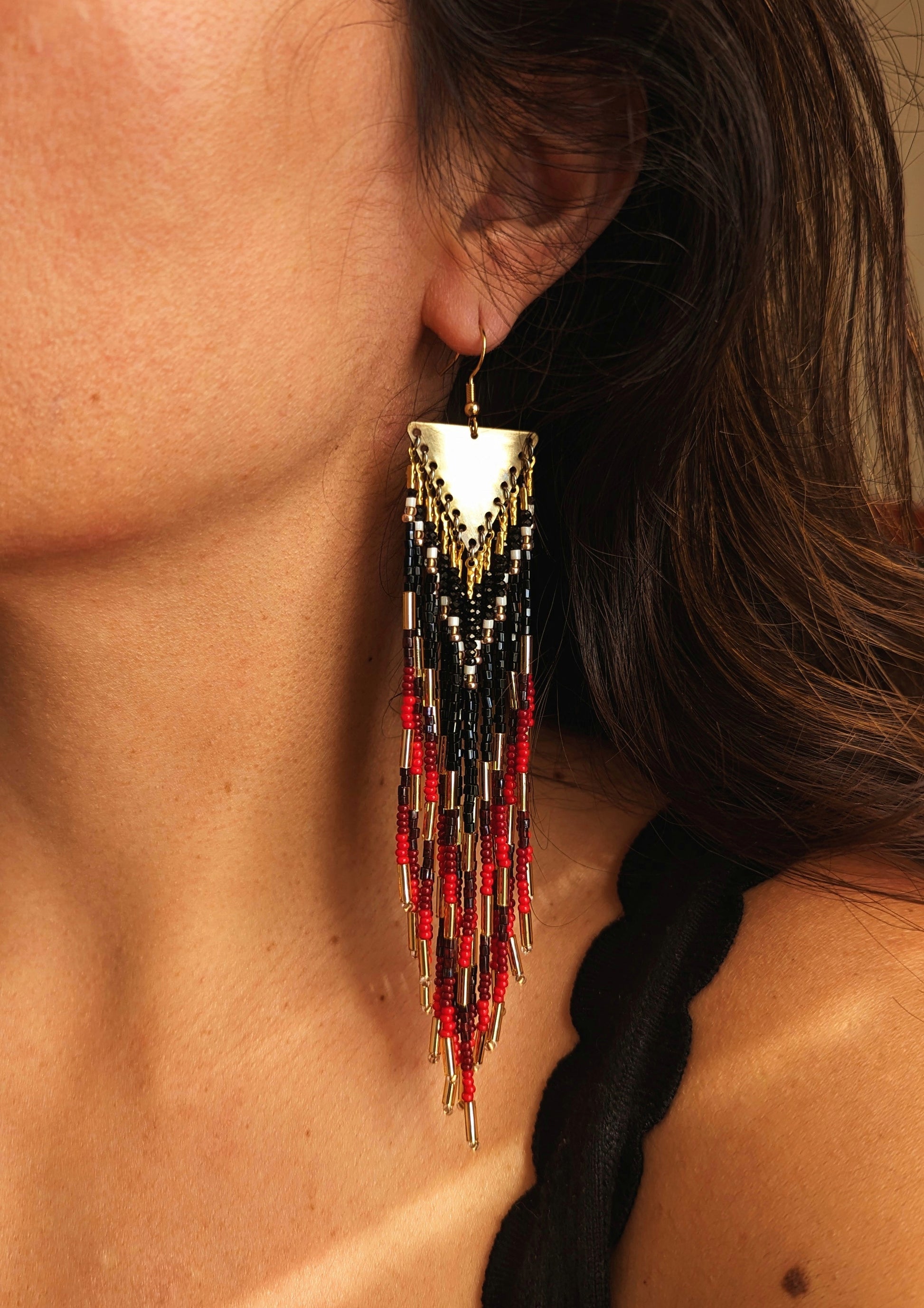 Moon & Milk earrings featuring black, red, and gold seed beads with brass triangle tops, worn by a woman.