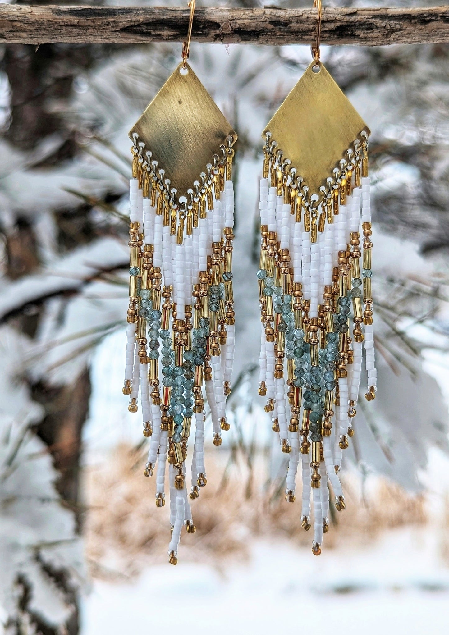 Beaded earrings with white, gold, and aqua tones hanging against a snowy winter backdrop, capturing a blend of elegance and nature.- Moon & Milk