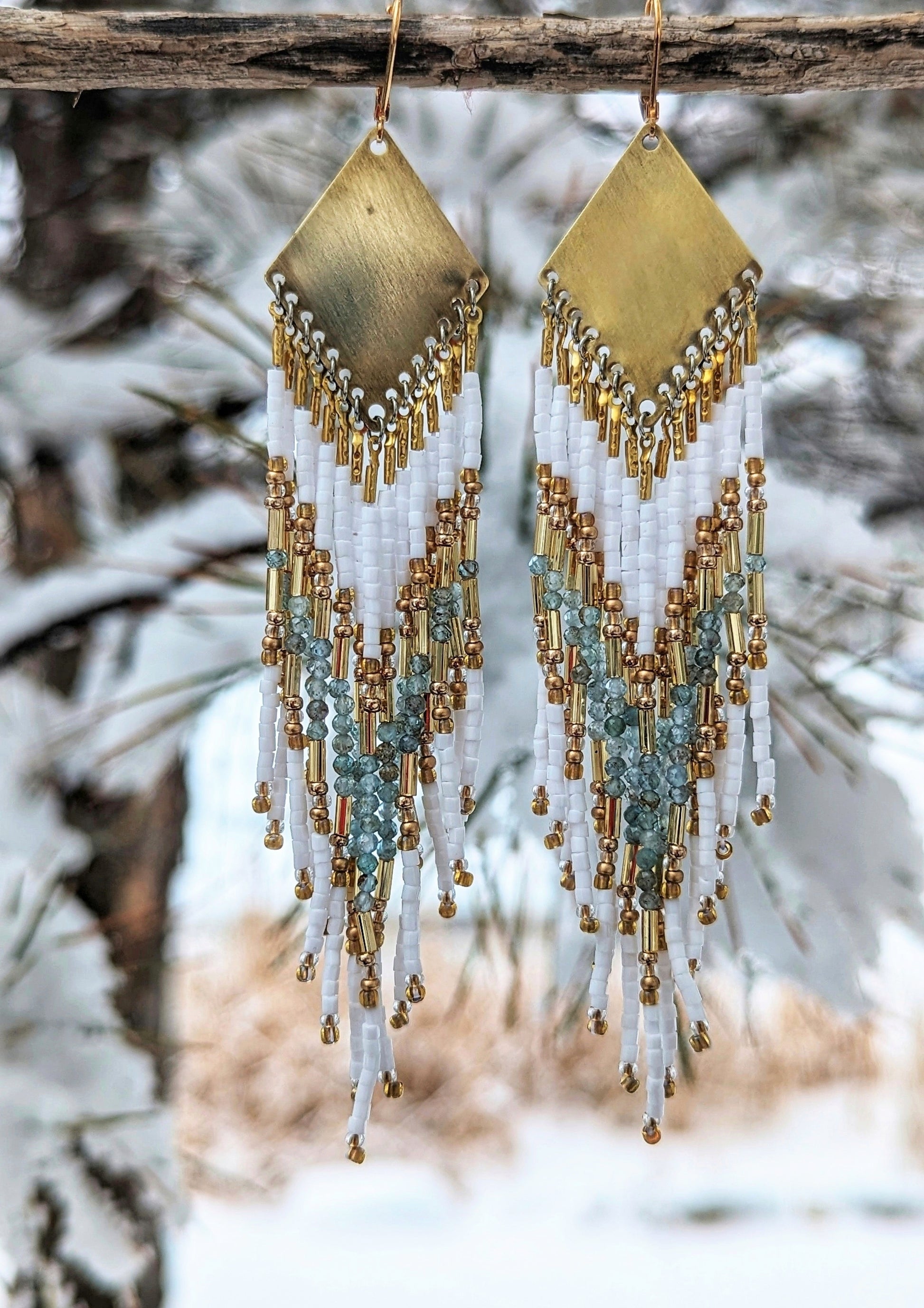 Beaded earrings with white, gold, and aqua tones hanging against a snowy winter backdrop, capturing a blend of elegance and nature.- Moon & Milk