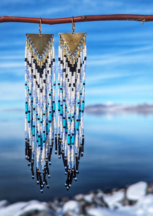 Moon & Milk - Long seed bead earrings with white, blue, black, and turquoise beads with a geometric  chevron pattern. Gold brass triangular charms. Hanging on a branch with a snowy background.