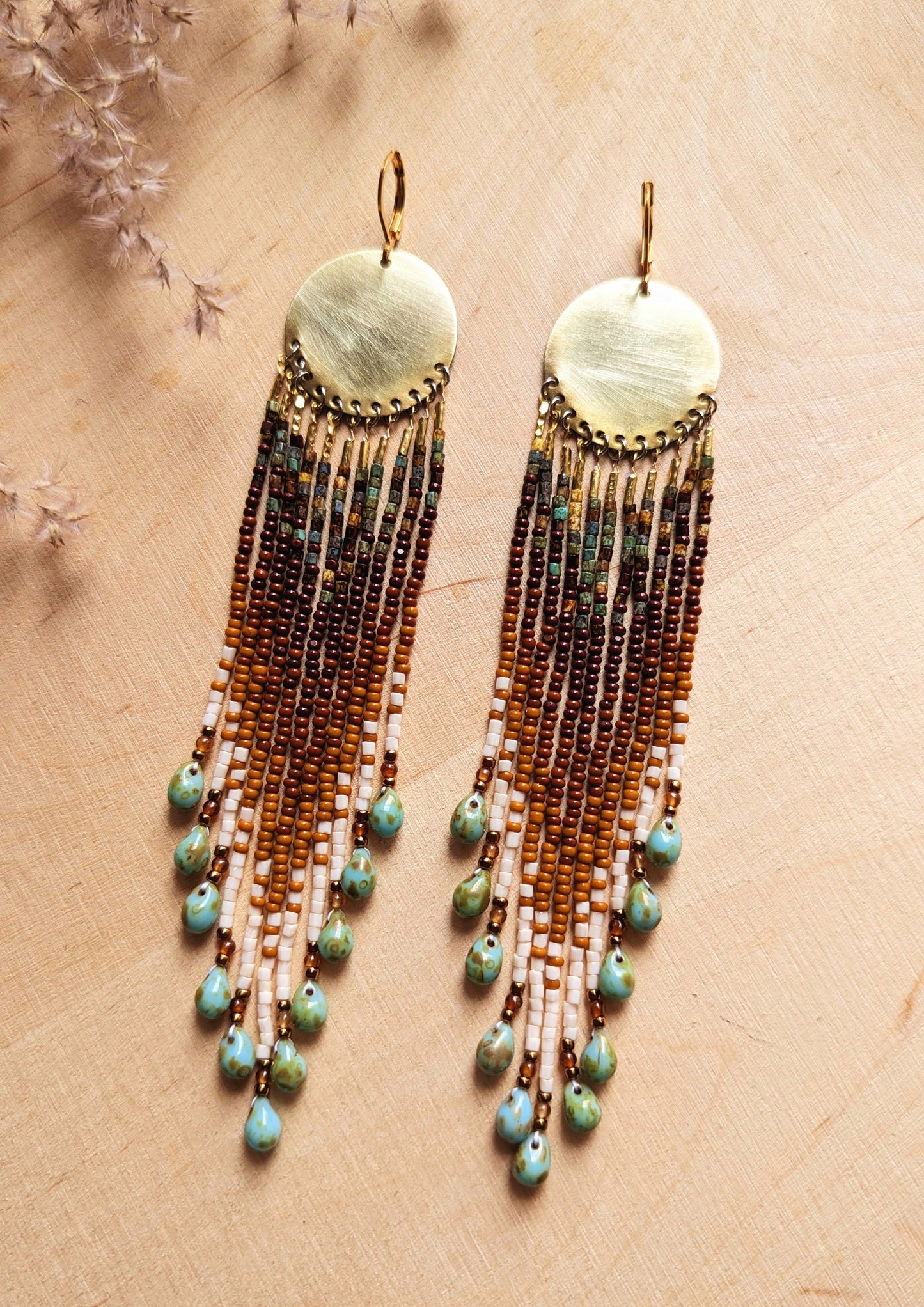 Pair of seed bead earrings with brass disks and long, earth-tone beaded fringe accented with turquoise teardrop beads, displayed on a wooden background with dried floral accents. - Moon & Milk
