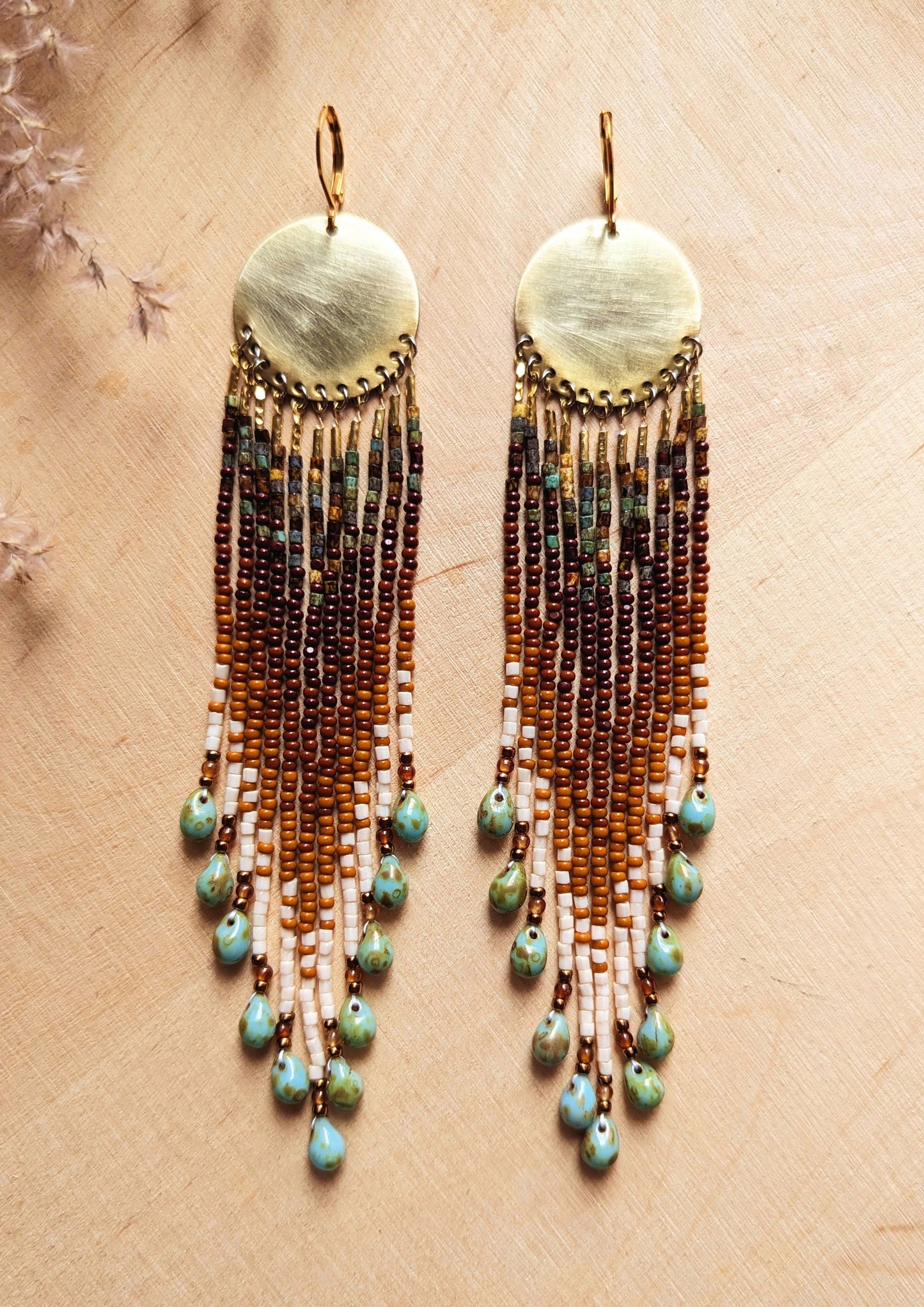 Pair of seed bead earrings with brass disks and long, earth-tone beaded fringe accented with turquoise teardrop beads, displayed on a wooden background with dried floral accents. - Moon & Milk