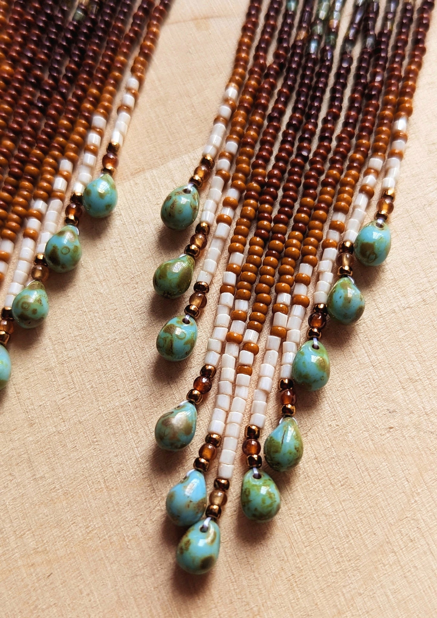 Close-up view of the fringe detail on earrings, showing brown, cream, and green beads, ending with small turquoise teardrop beads on a wooden surface. - Moon & Milk