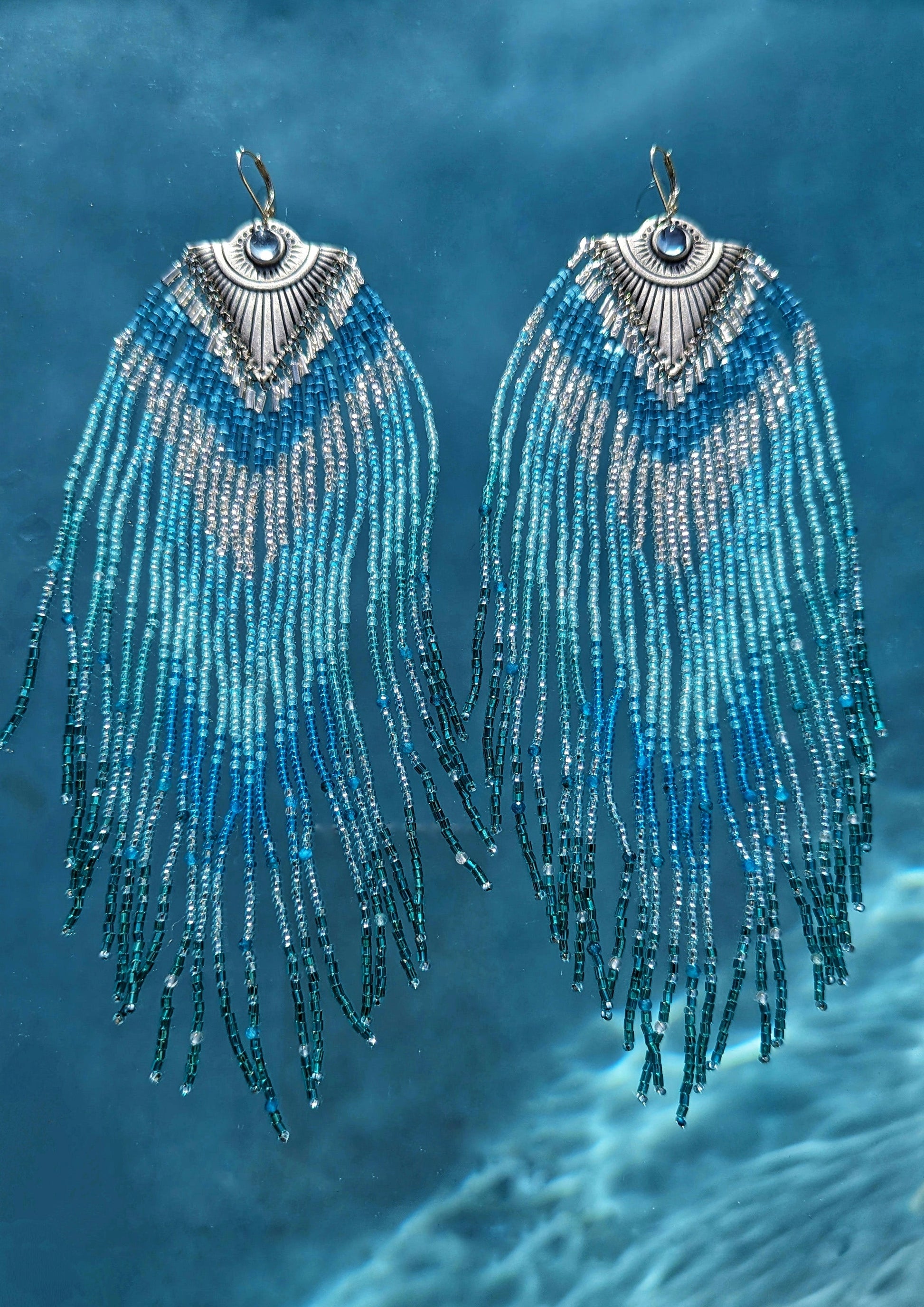 Moon & Milk - Stunning handmade beaded earrings with cascading blue and turquoise beads, featuring a silver-tone pendant with a central blue gemstone, elegantly displayed against a background of clear blue water.
