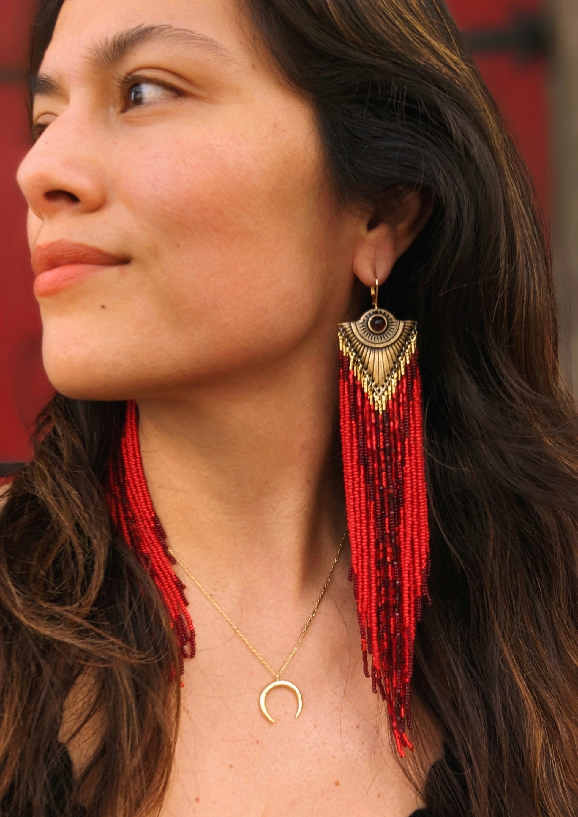 Moon & Milk - Woman wearing long, dangling red seed bead earrings with sparkly and gold beads, featuring an ornate gold metal top. She is also wearing a gold crescent moon necklace.