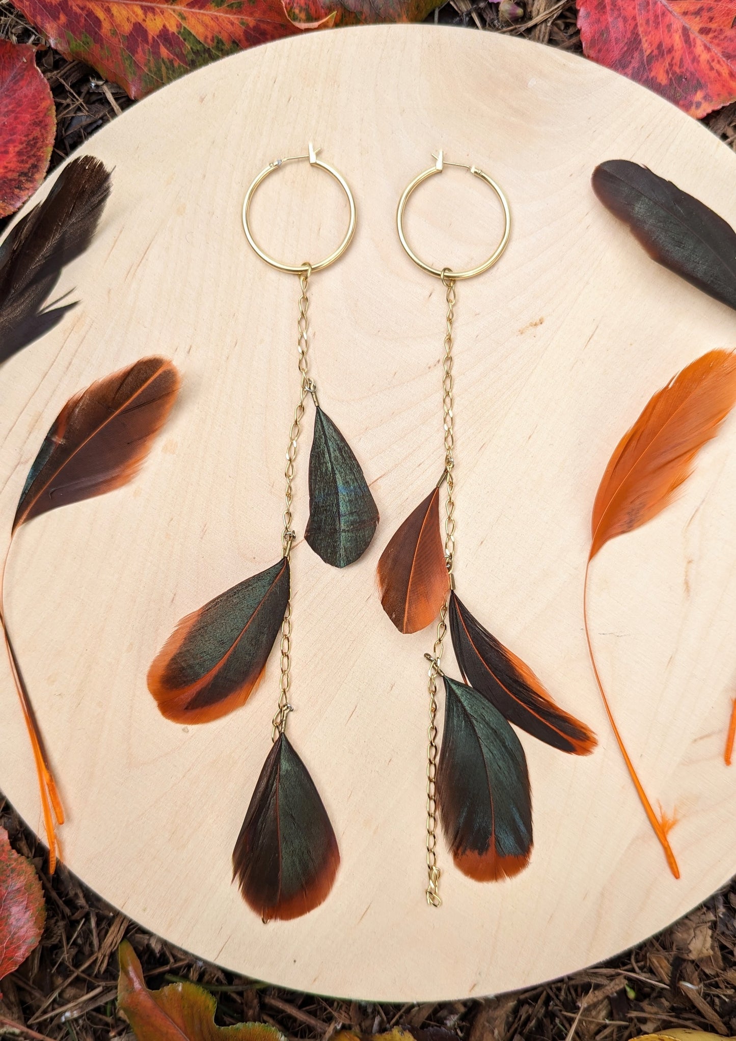 Boho feather earrings by Moon & Milk featuring gold hoops with cascading chains and feathers in dark green and orange tones, photographed with autumn leaves.