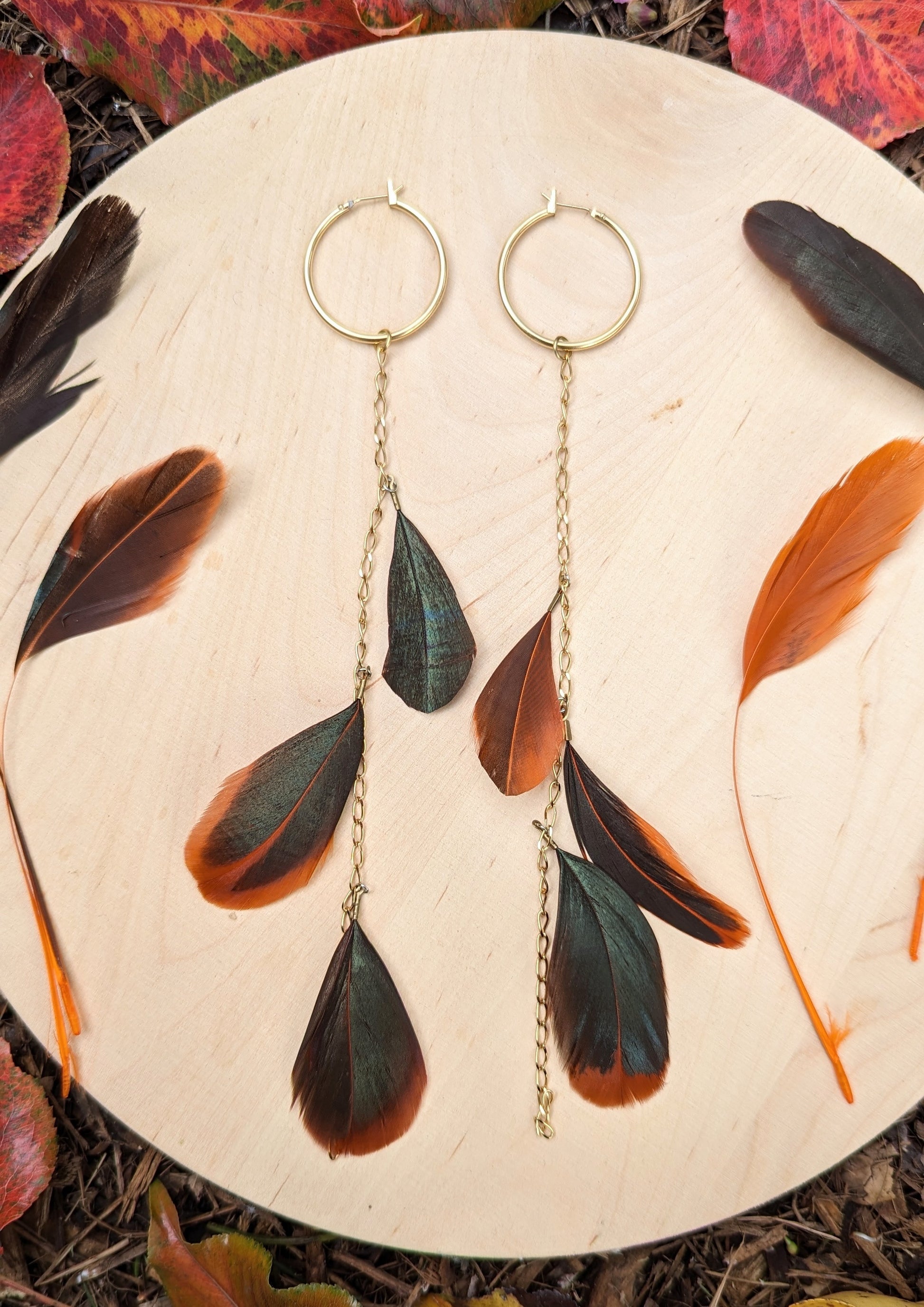 Boho feather earrings by Moon & Milk featuring gold hoops with cascading chains and feathers in dark green and orange tones, photographed with autumn leaves.