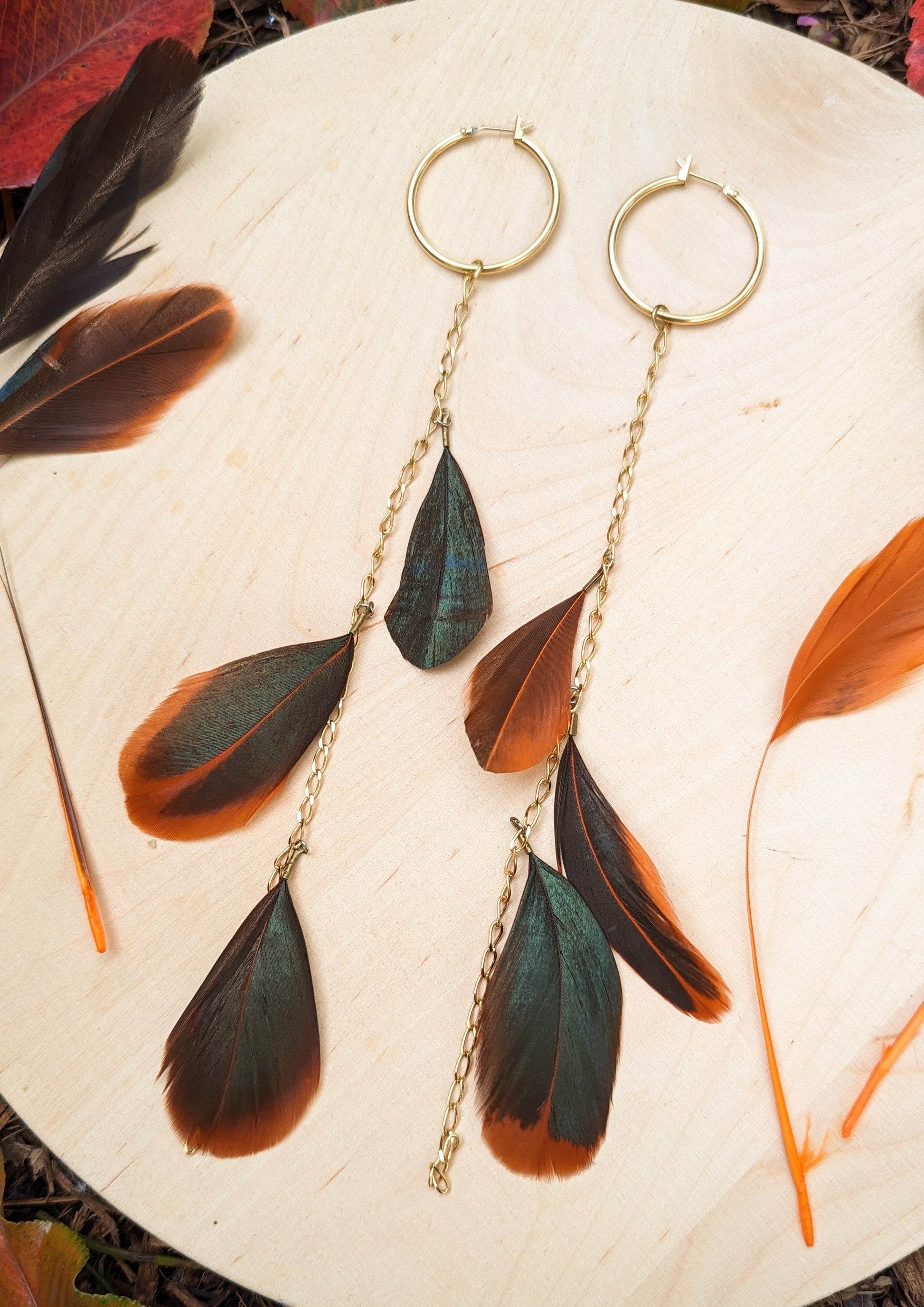 Boho feather earrings by Moon & Milk featuring gold hoops with cascading chains and feathers in dark green and orange tones, photographed with autumn leaves.