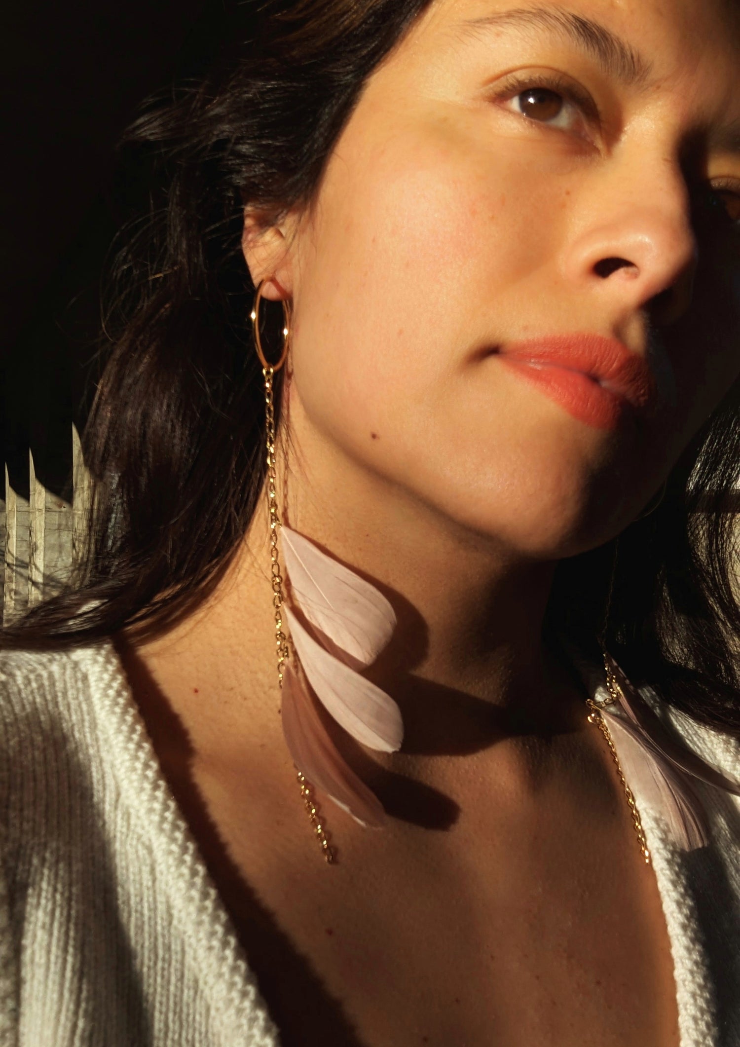 Close-up of a woman wearing Moon & Milk’s blush pink feather earrings with gold chain detail, glowing in golden-hour light.