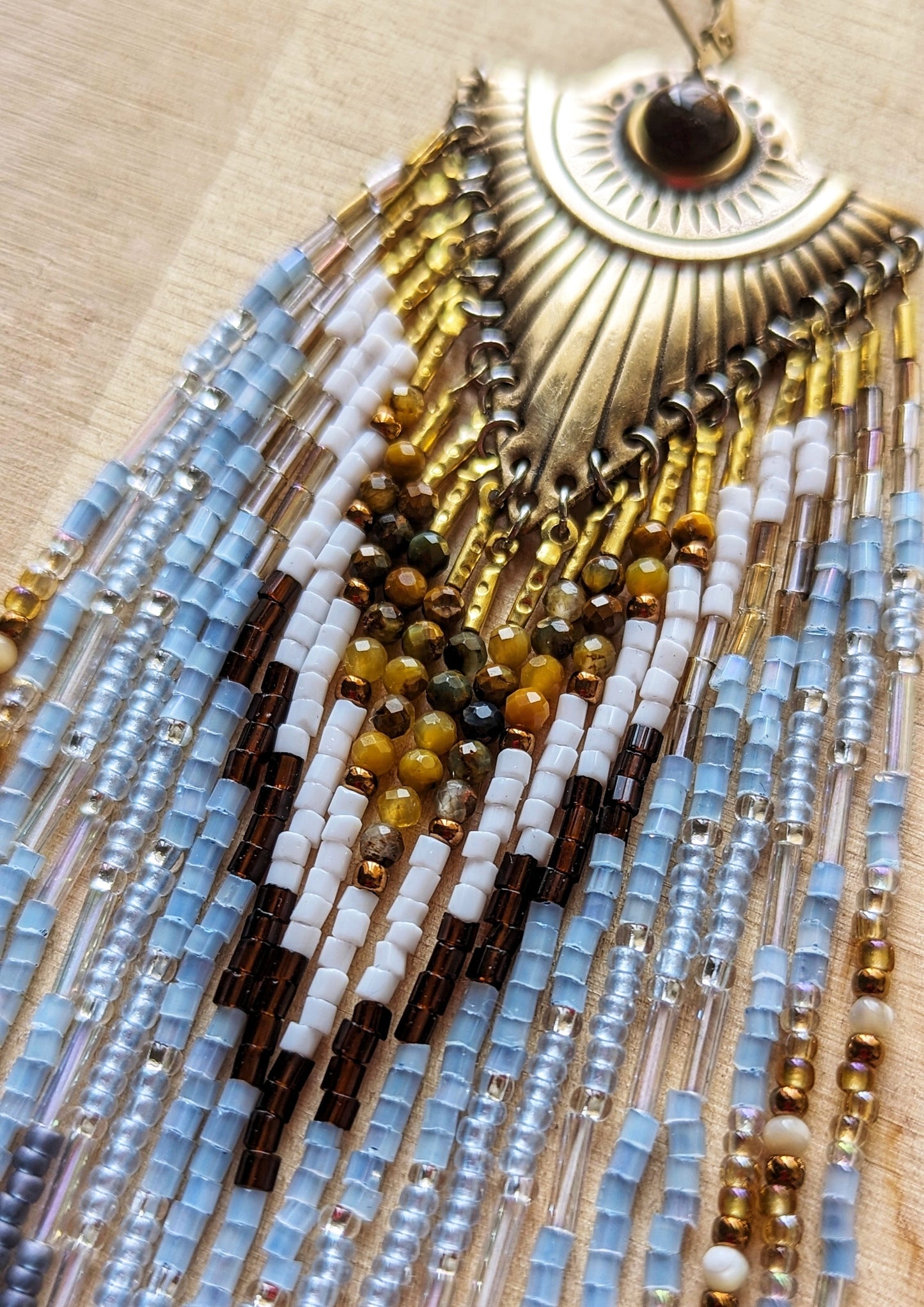 Long dangling seed bead earrings with white, blue, and gold beads in a chevron pattern. Ornate gold metal tops. Displayed on a wooden surface. Statement piece.
