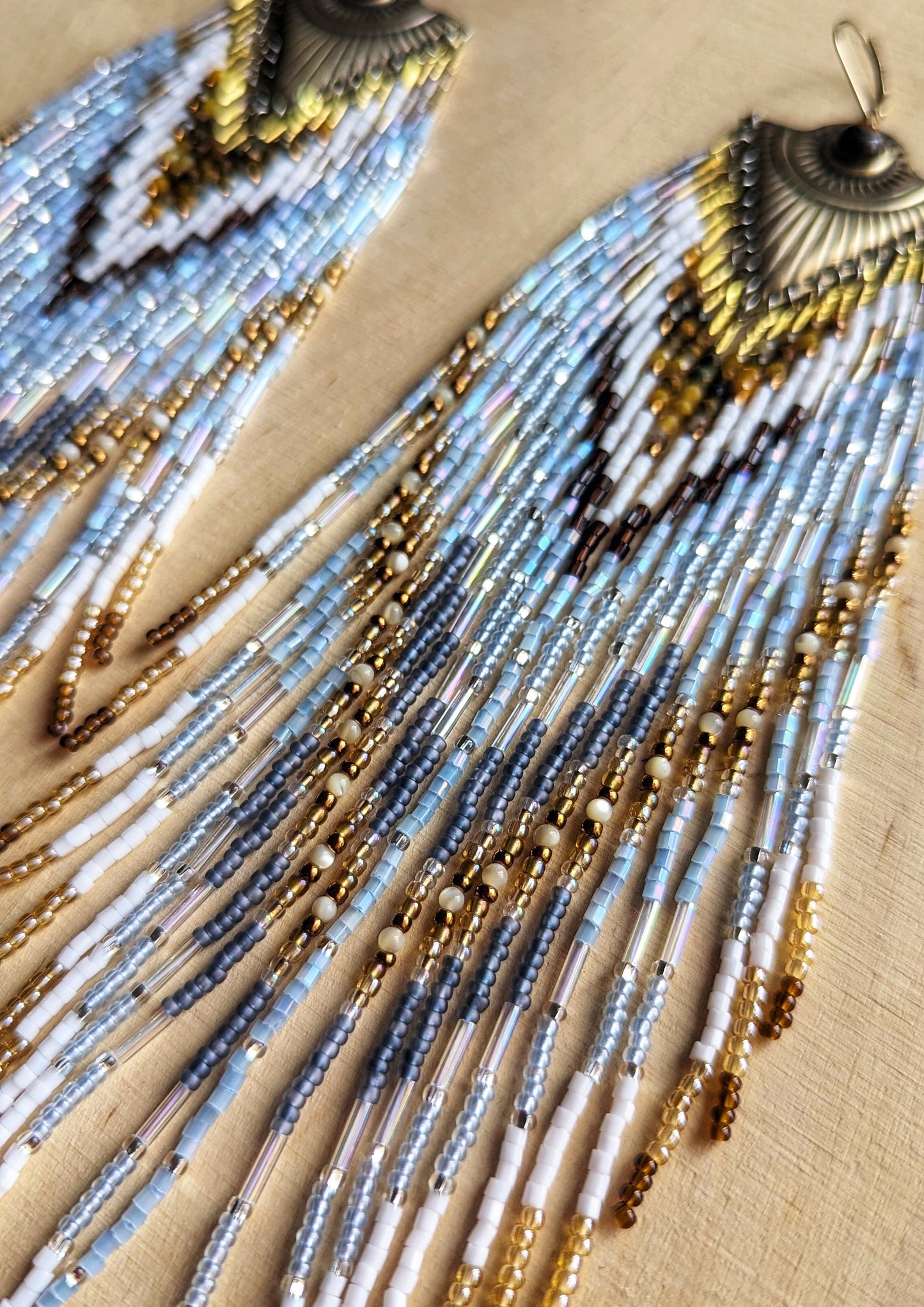 Long dangling seed bead earrings with white, blue, and gold beads in a chevron pattern. Ornate gold metal tops. Displayed on a wooden surface. Statement piece.