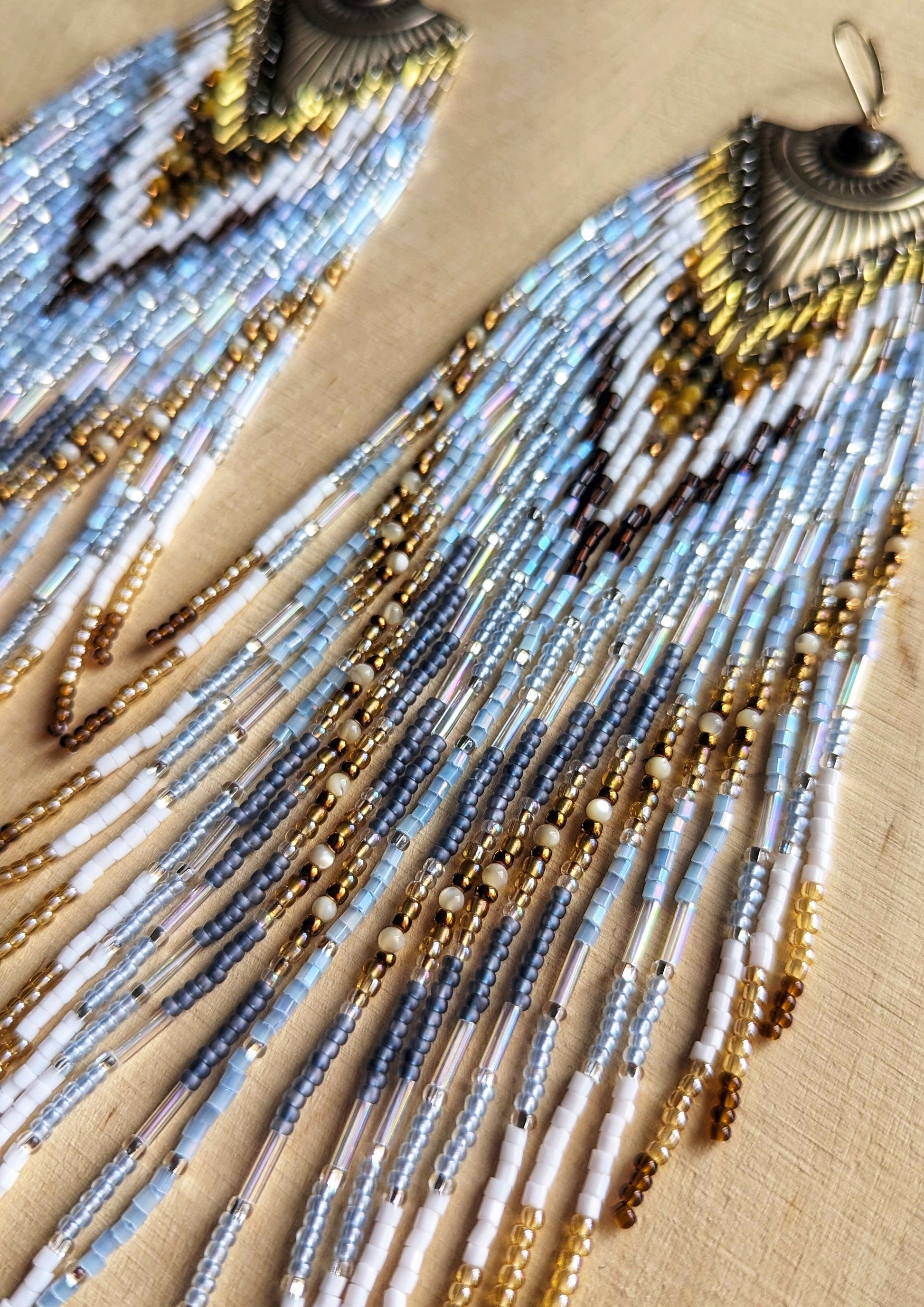 Long dangling seed bead earrings with white, blue, and gold beads in a chevron pattern. Ornate gold metal tops. Displayed on a wooden surface. Statement piece.