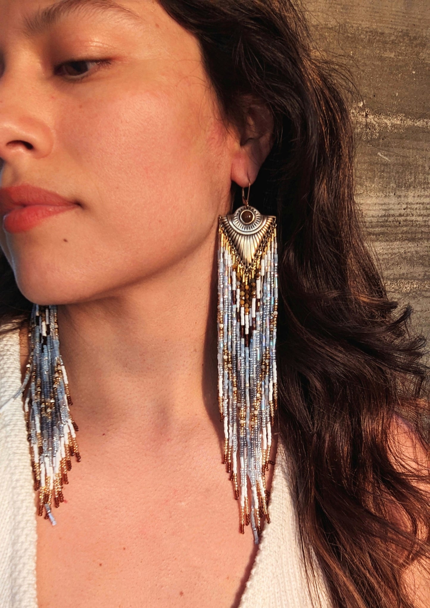 Woman wearing long dangling seed bead earrings with white, blue, and gold beads, featuring an ornate gold metal top. She has long dark hair.